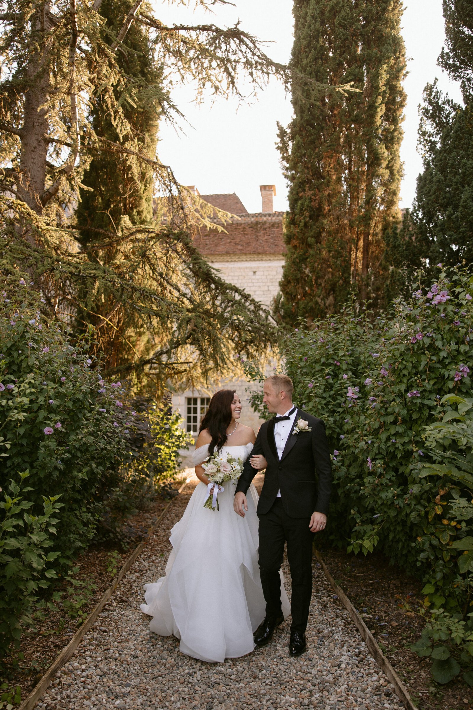 Bride and groom walking down a gravel path, surrounded by greenery and tall trees, with a building in the background.