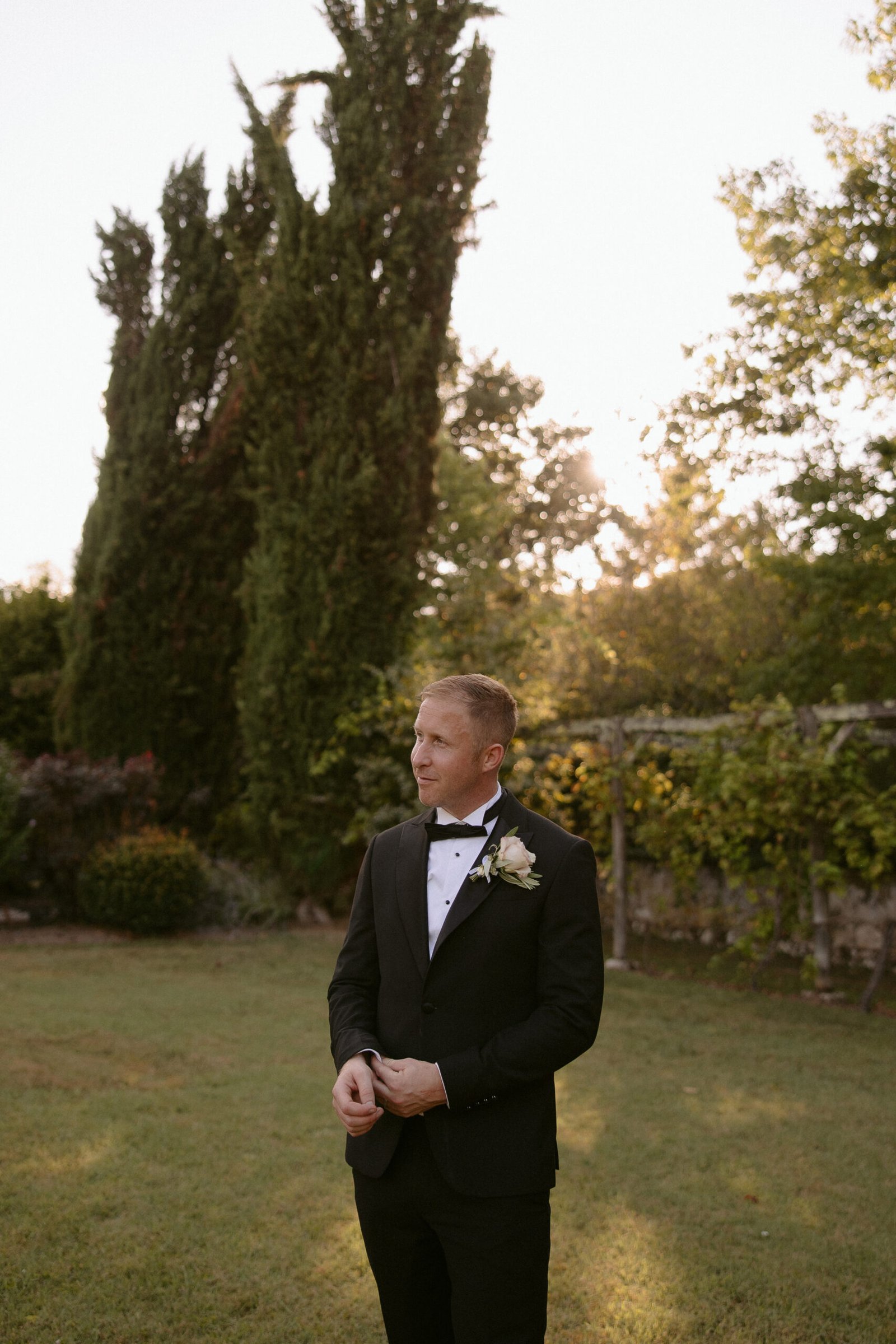 Man in a tuxedo stands on grass in a garden, adjusting his cufflinks. Tall trees and greenery are in the background, with sunlight filtering through.