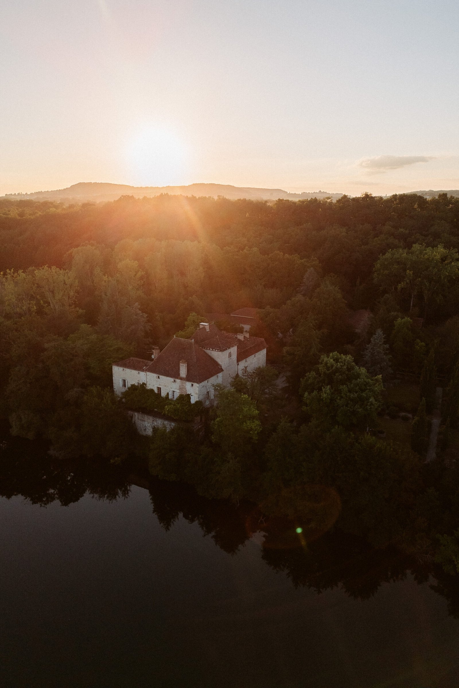 Sunset over a landscape with a forest-bordered building near a calm body of water, and distant hills on the horizon.