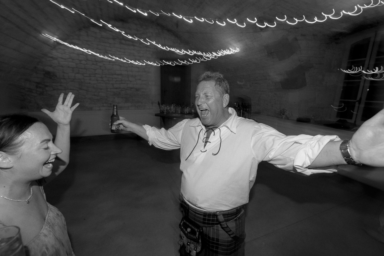 A man joyfully sings with arms outstretched, holding a drink, while a woman with raised hands faces him in a blurred, festive room with string lights.