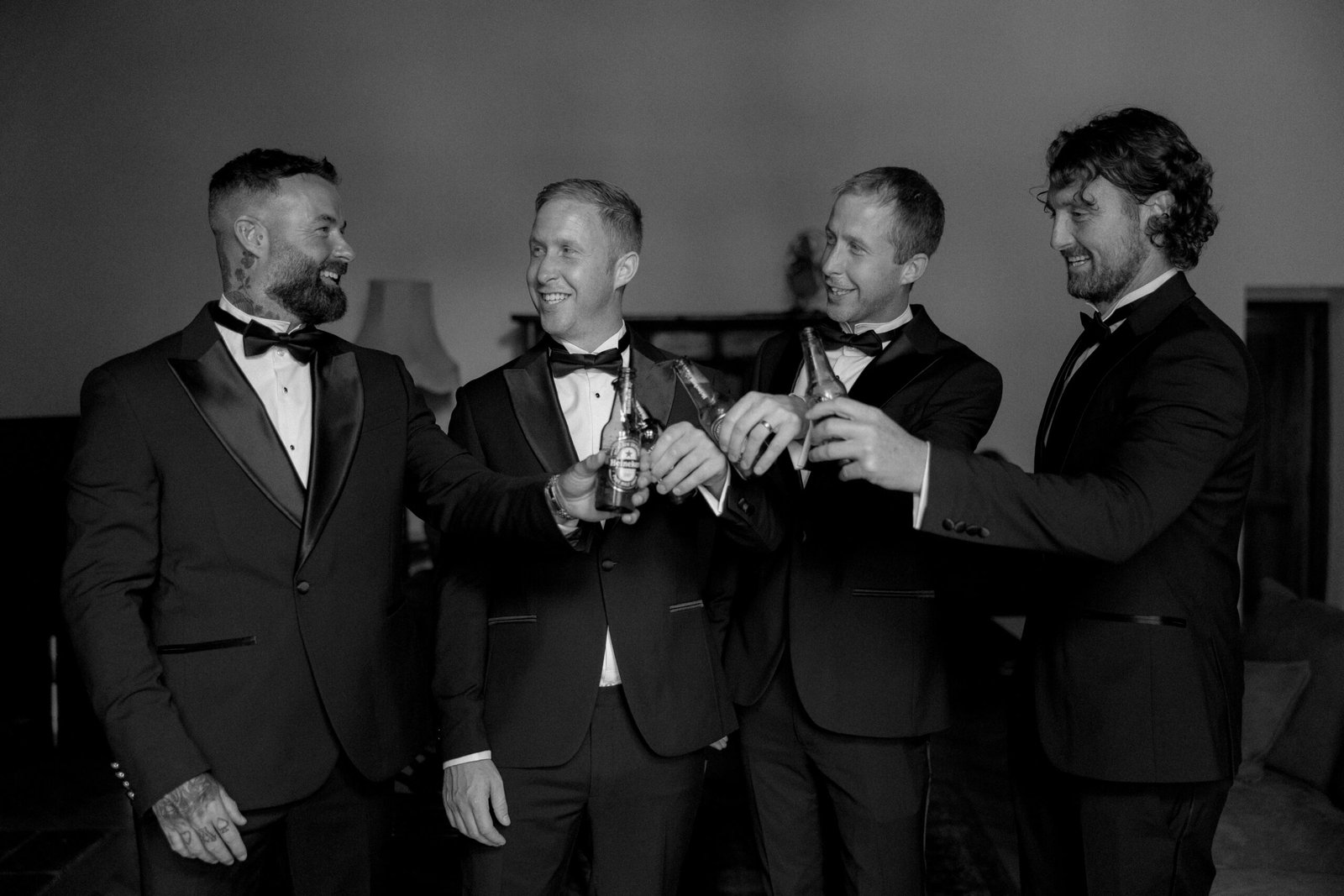 Four men in tuxedos toast with drinks, smiling and standing together indoors.