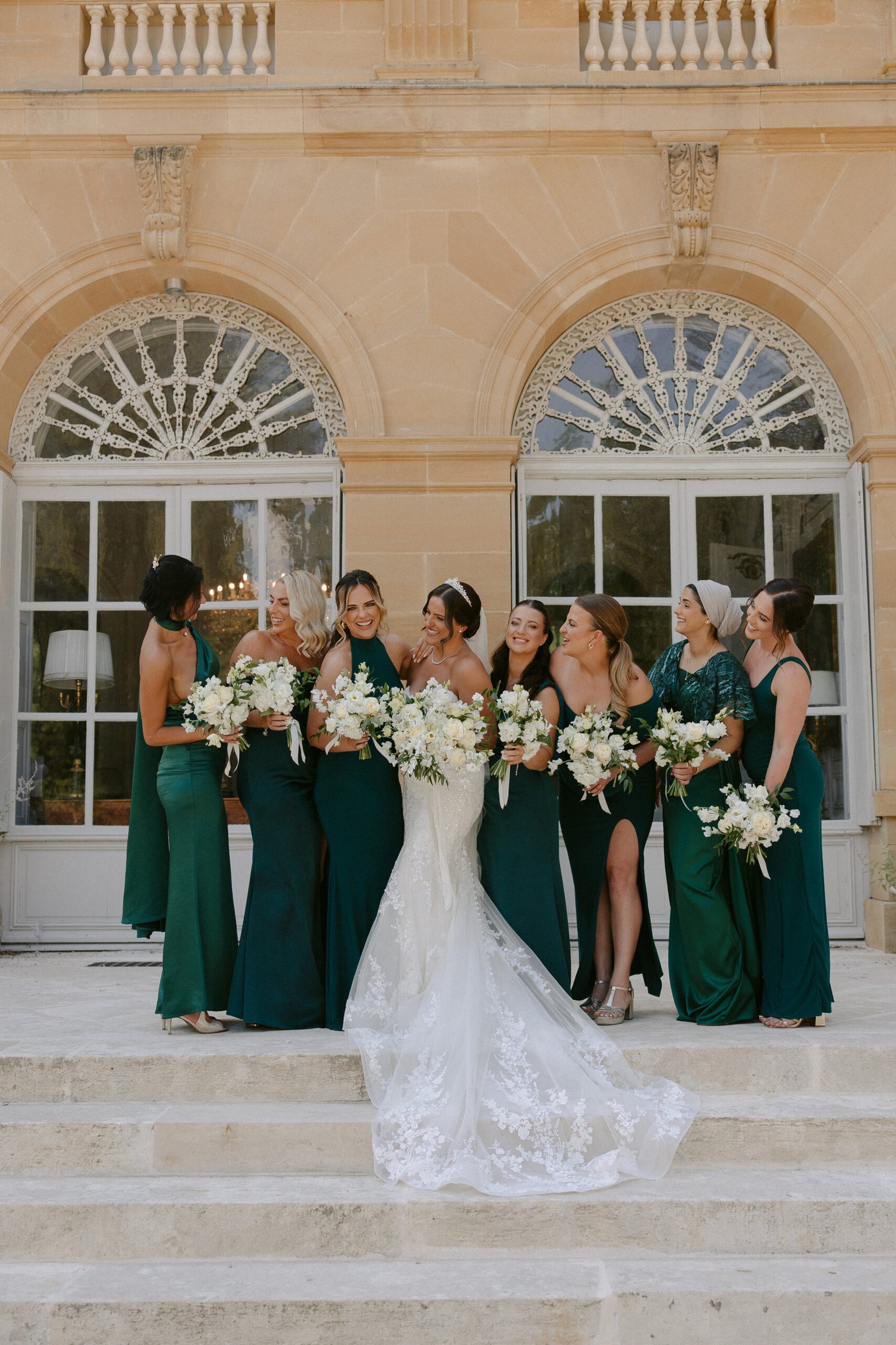 A bride in a white dress stands with her bridesmaids in green dresses, holding bouquets, on the steps of a large building with arched windows. Chateau la Durantie Wedding Photographer.