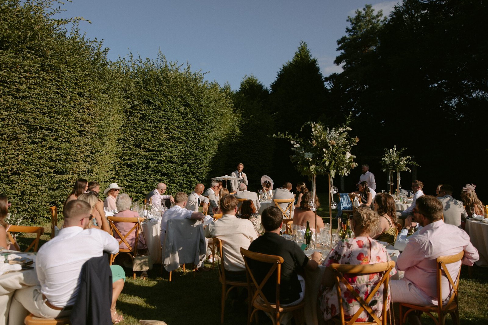Outdoor garden event with seated guests at tables, surrounded by tall hedges. A person at the front appears to be speaking. Sunny weather and clear skies. Chateau la Durantie Wedding Photographer.