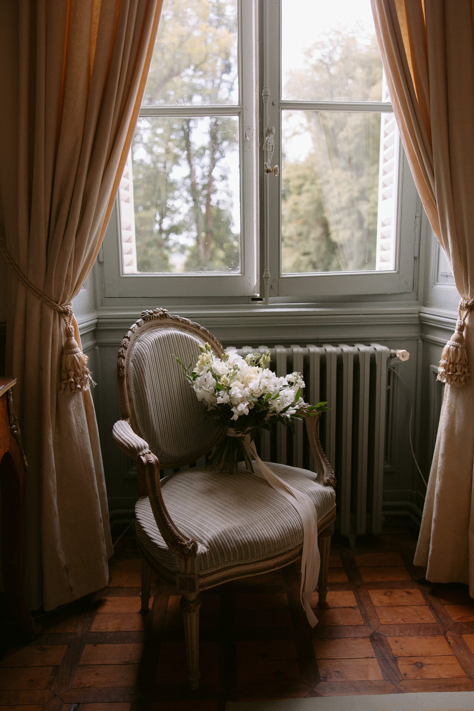 A vintage armchair with a bouquet of white flowers is placed in front of a window with beige curtains, overlooking a view of trees.