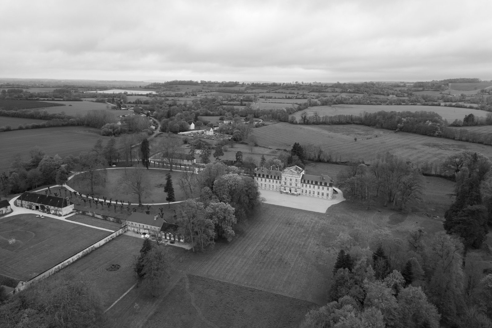 Aerial view of a large historic estate with a main building, courtyard, surrounding trees, and fields under a cloudy sky.