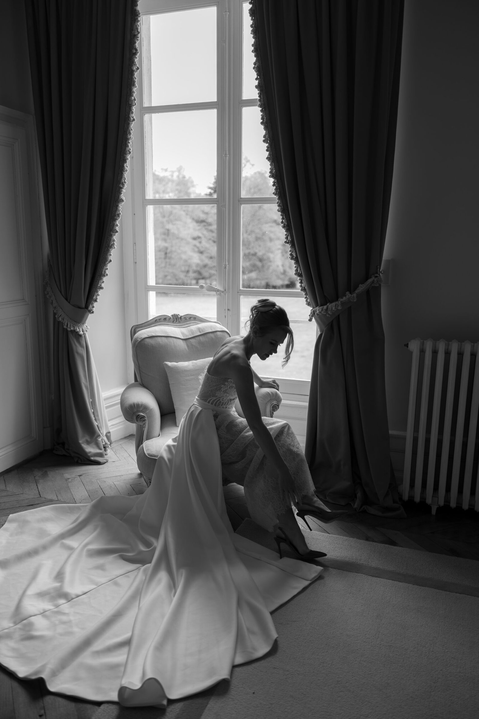A woman in a bridal gown sits on a chair near a large window with curtains, adjusting her shoe in a softly lit room.