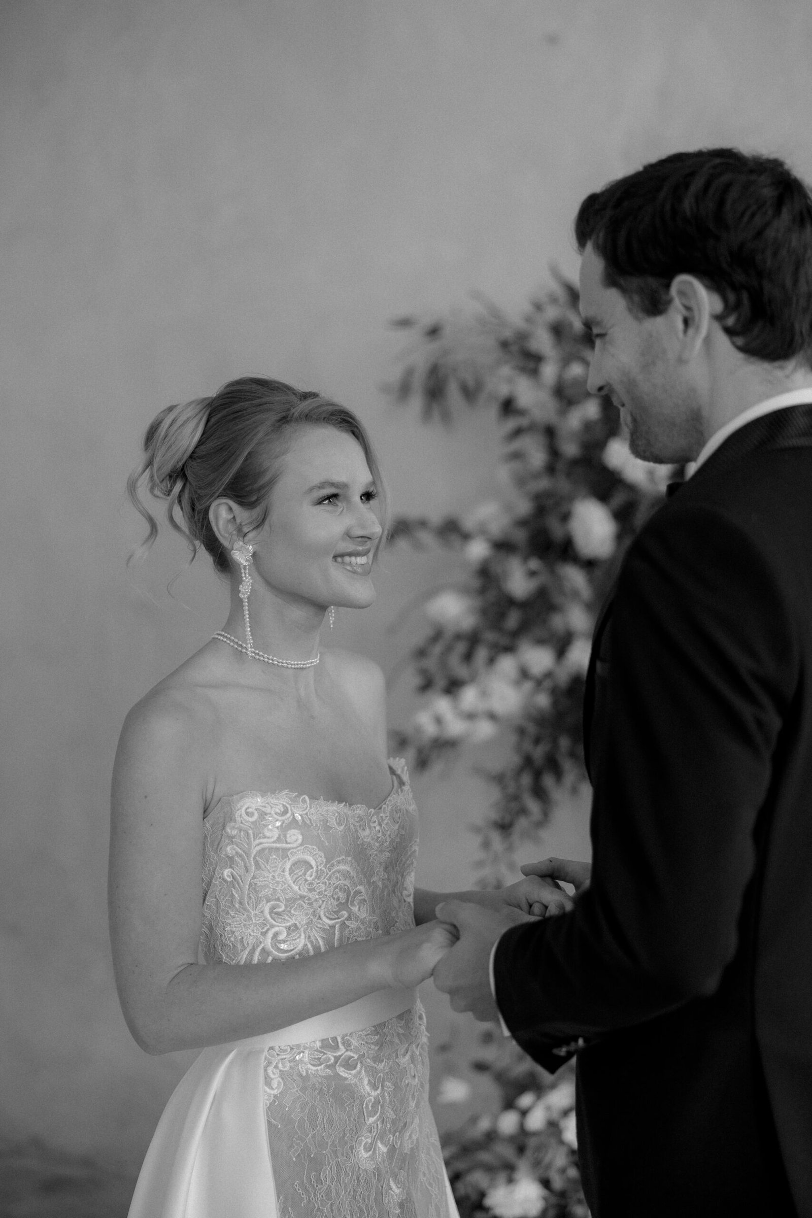 A bride and groom stand facing each other holding hands, dressed in wedding attire, with floral arrangements in the background.