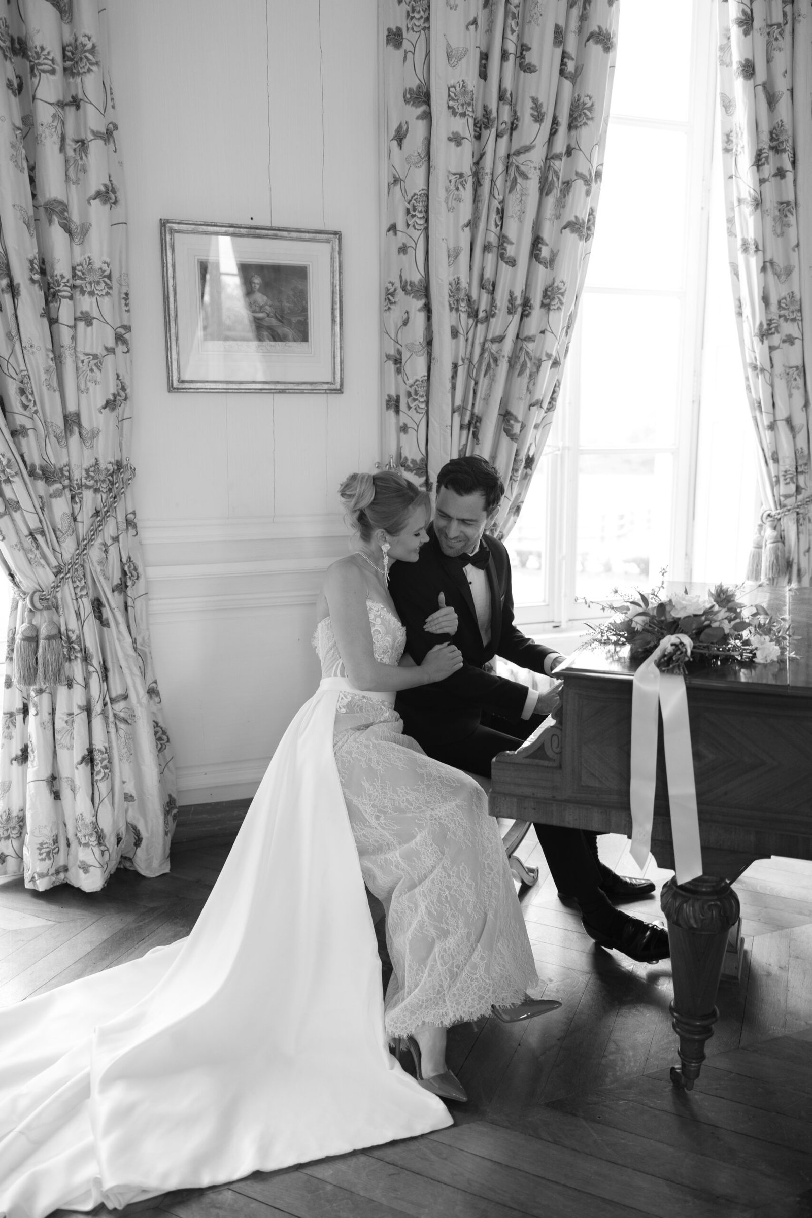 A bride and groom sit together at a piano near a window, dressed in formal wedding attire, with a bouquet on the piano. Chateau de Courtomer wedding photographer.