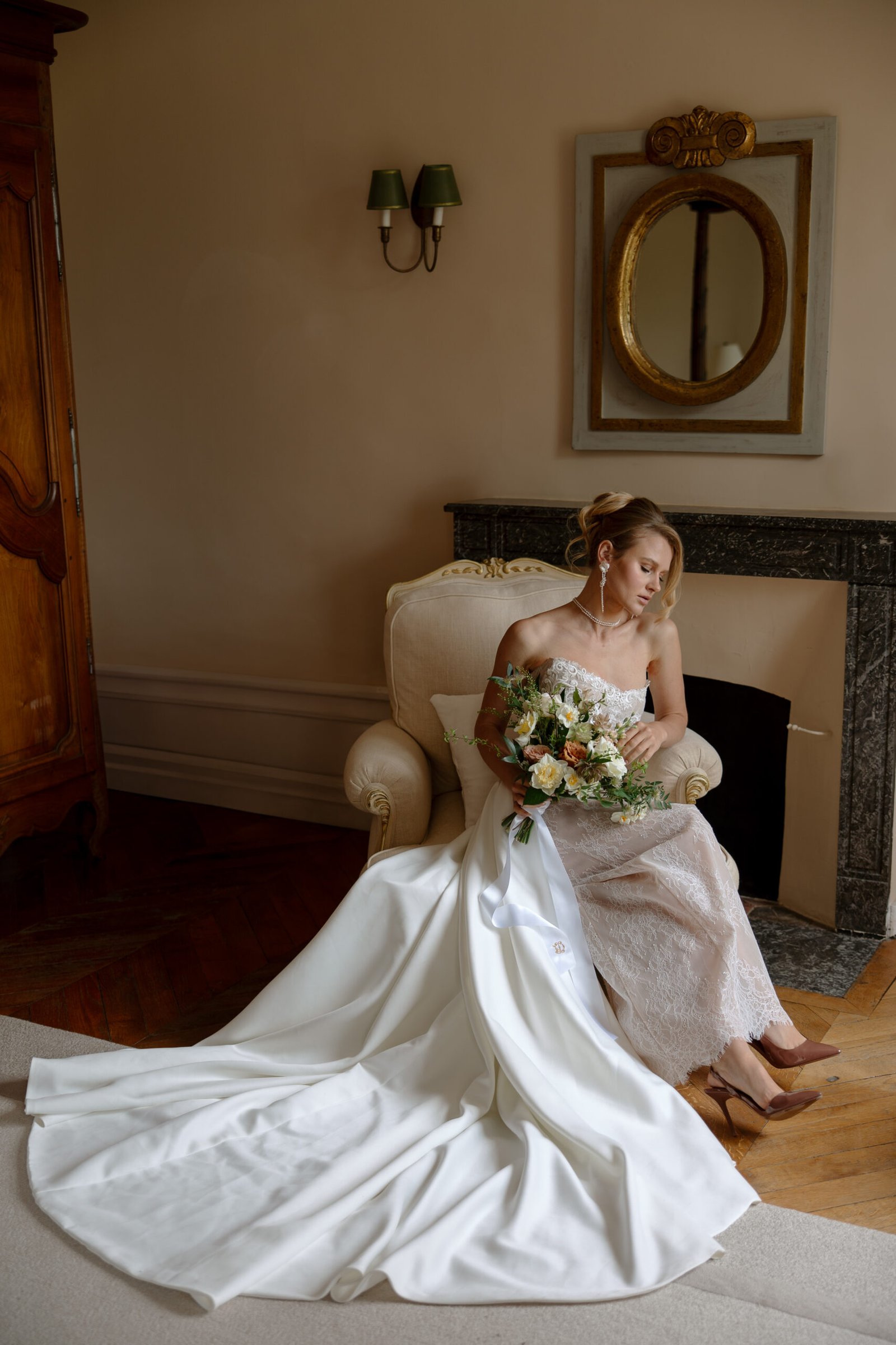 A woman in a white wedding dress sits on an armchair, holding a bouquet of flowers, next to a fireplace with a round mirror above it.