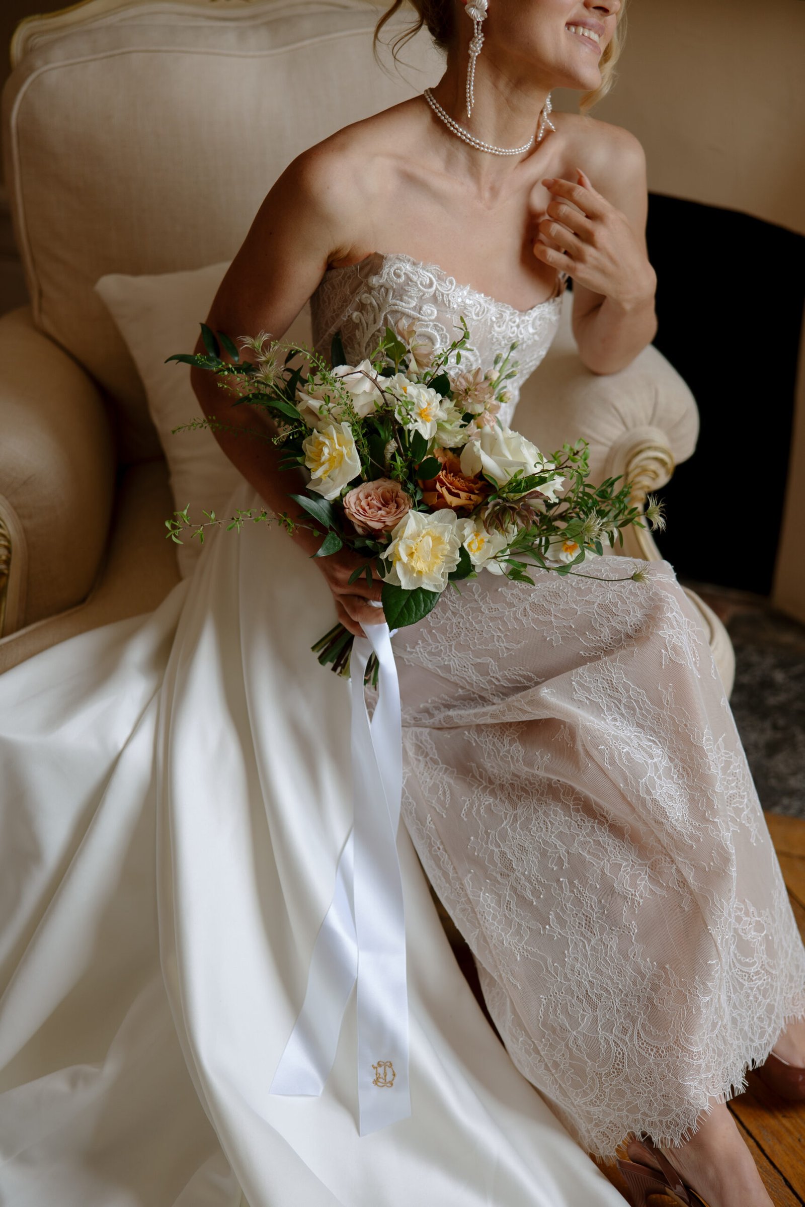 A bride in a strapless lace wedding dress sits on a beige armchair, holding a bouquet of flowers with a white ribbon.