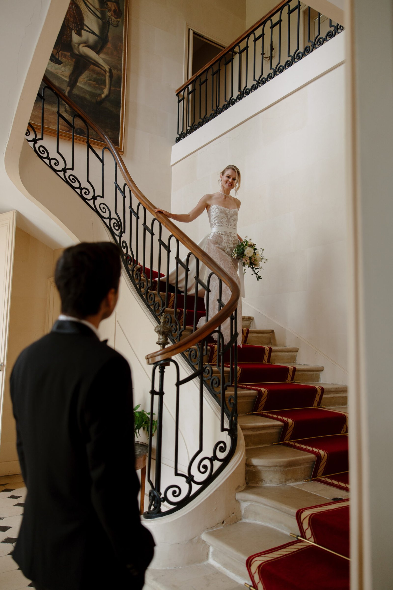 A bride in a white dress holding a bouquet descends a curved staircase, while a man in a black suit looks up at her.
