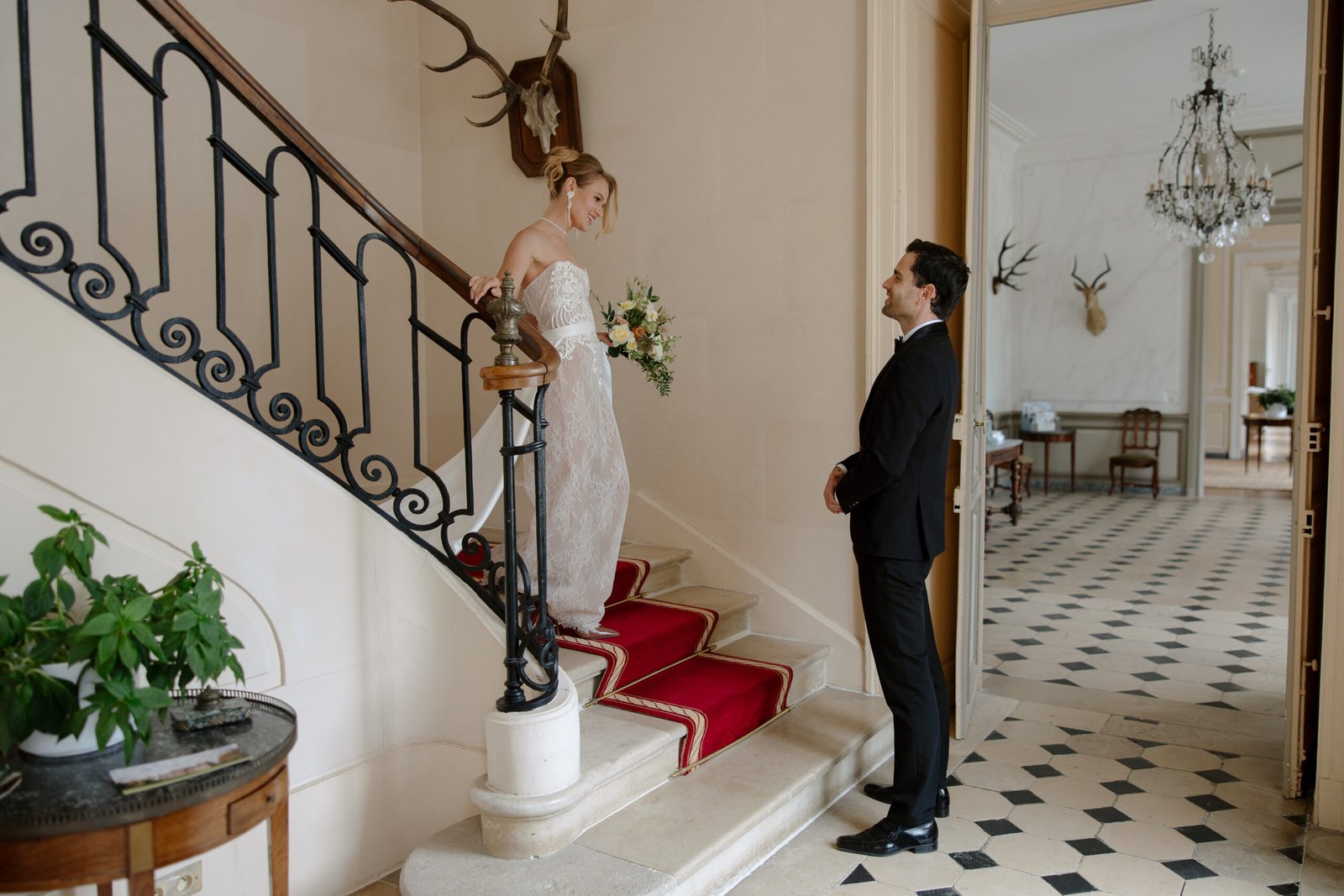 A bride in a white dress descends a staircase toward a groom in a black suit, inside an elegant room with tiled floors and antler decor.