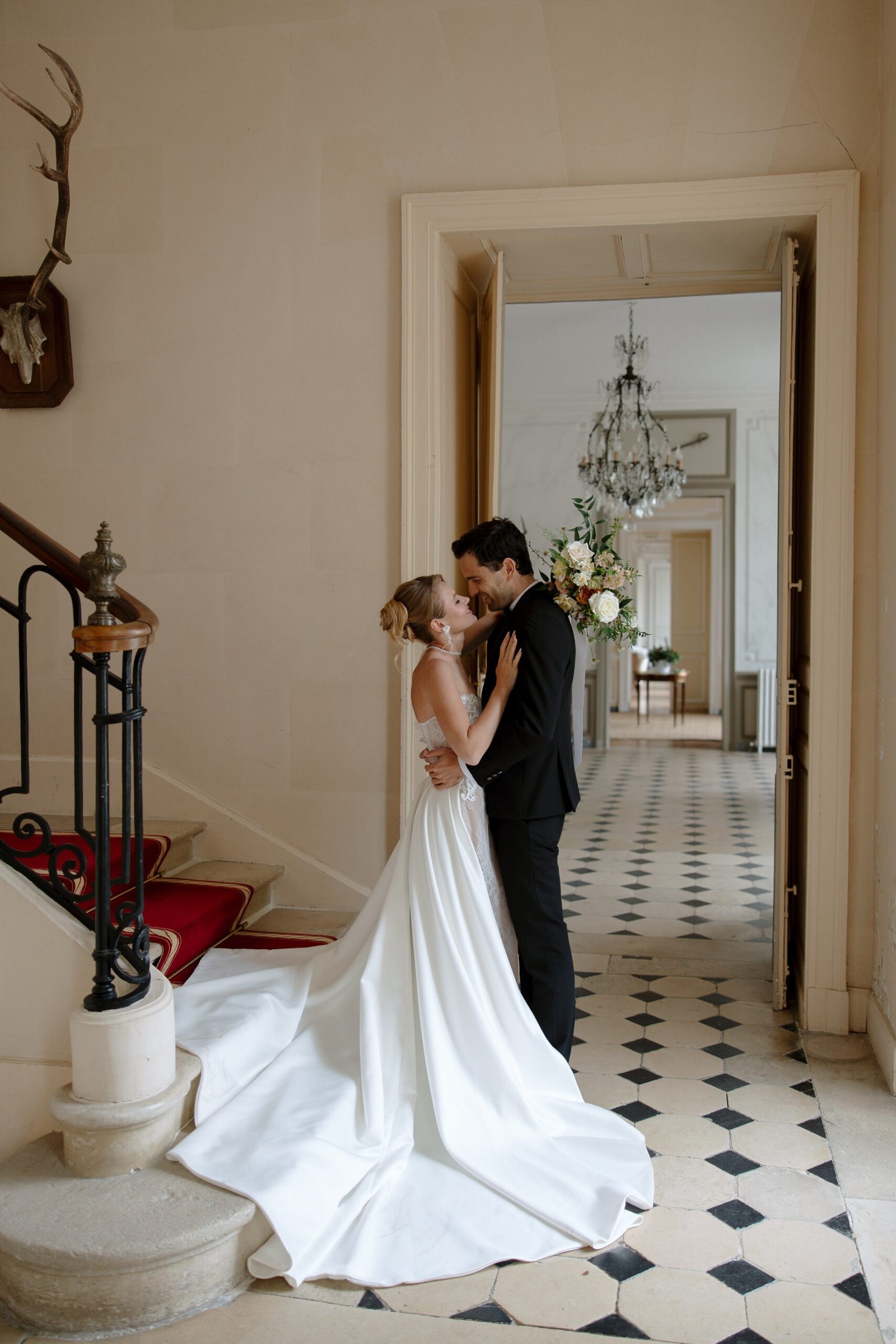 A bride and groom embrace on a black-and-white tiled floor in an elegant hallway with a chandelier and antlers mounted on the wall.