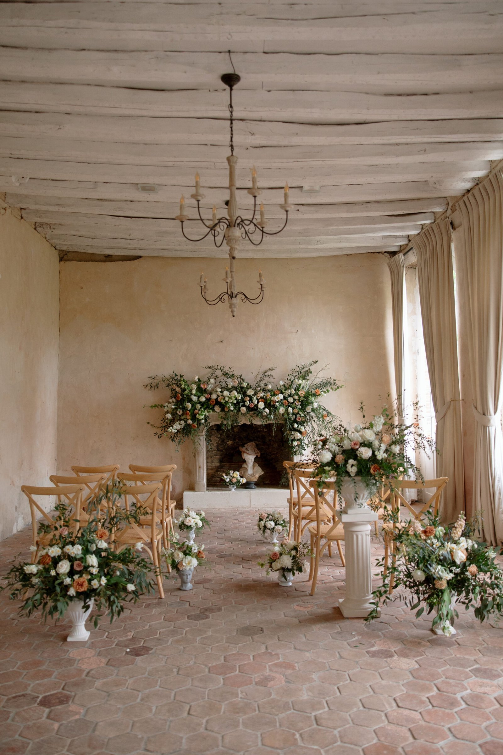 A small indoor wedding ceremony setup features wooden chairs, floral arrangements, a rustic chandelier, a brick floor, and a fireplace decorated with flowers.