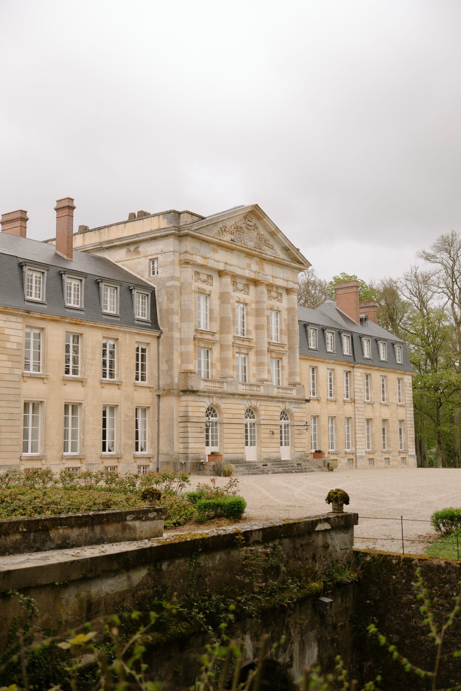 A large, beige stone chateau with tall windows and a gravel driveway, surrounded by greenery under an overcast sky.