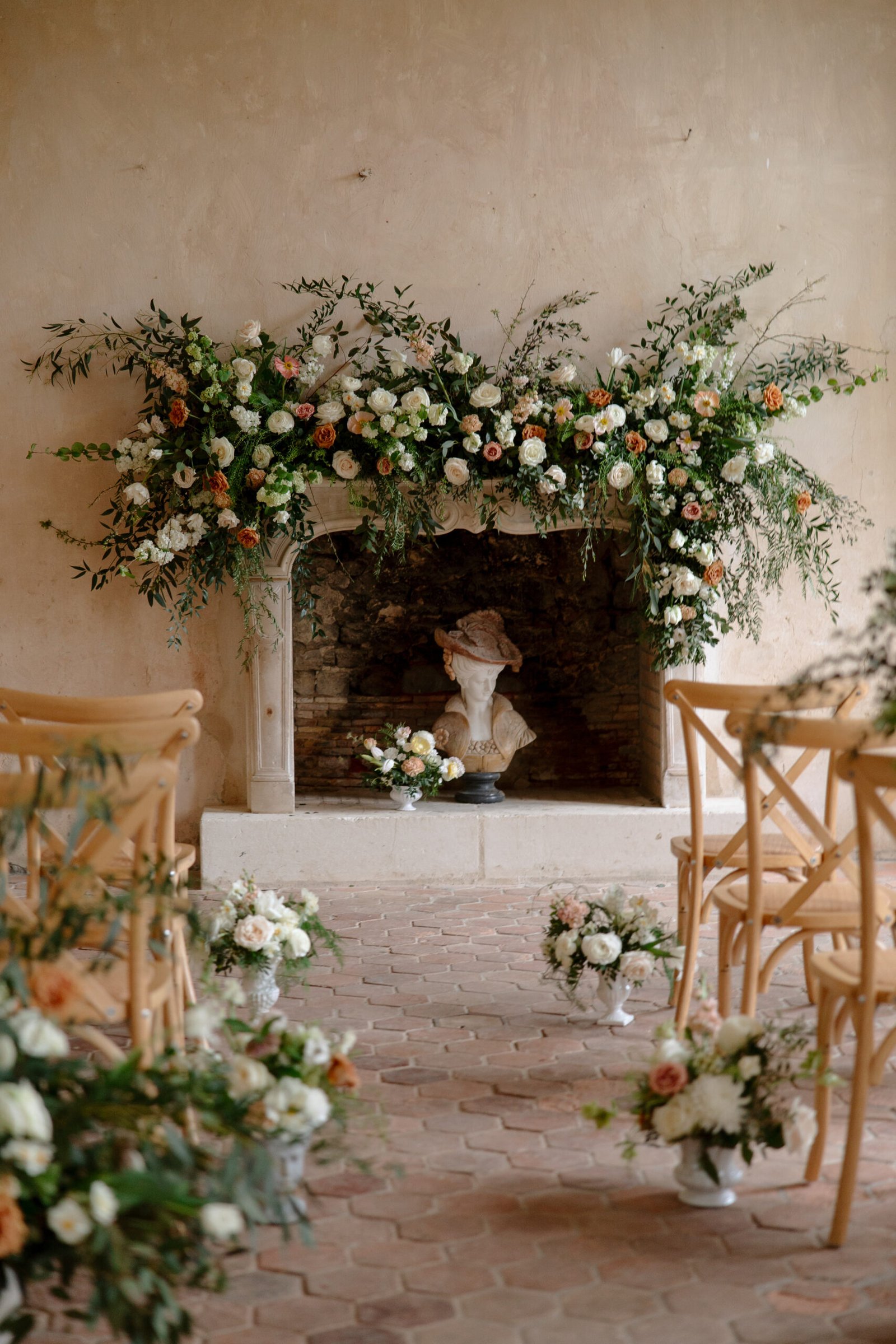 Floral arrangements with white and peach flowers decorate a stone fireplace and aisle, with wooden chairs set up for an indoor ceremony.