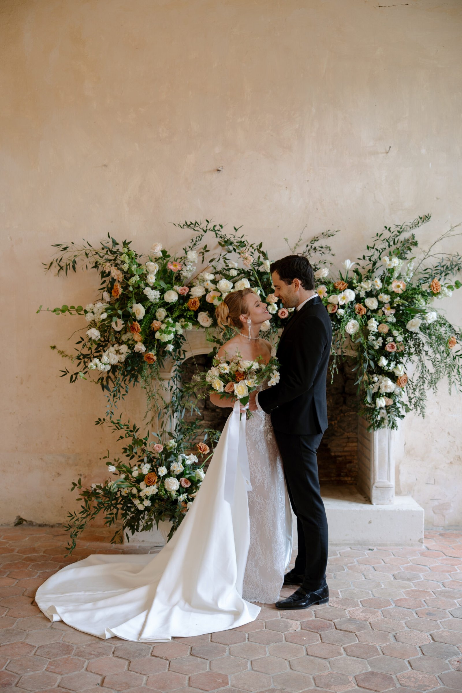 A bride and groom stand together in front of a floral arrangement, facing each other and holding a bouquet, with a neutral-toned wall in the background.