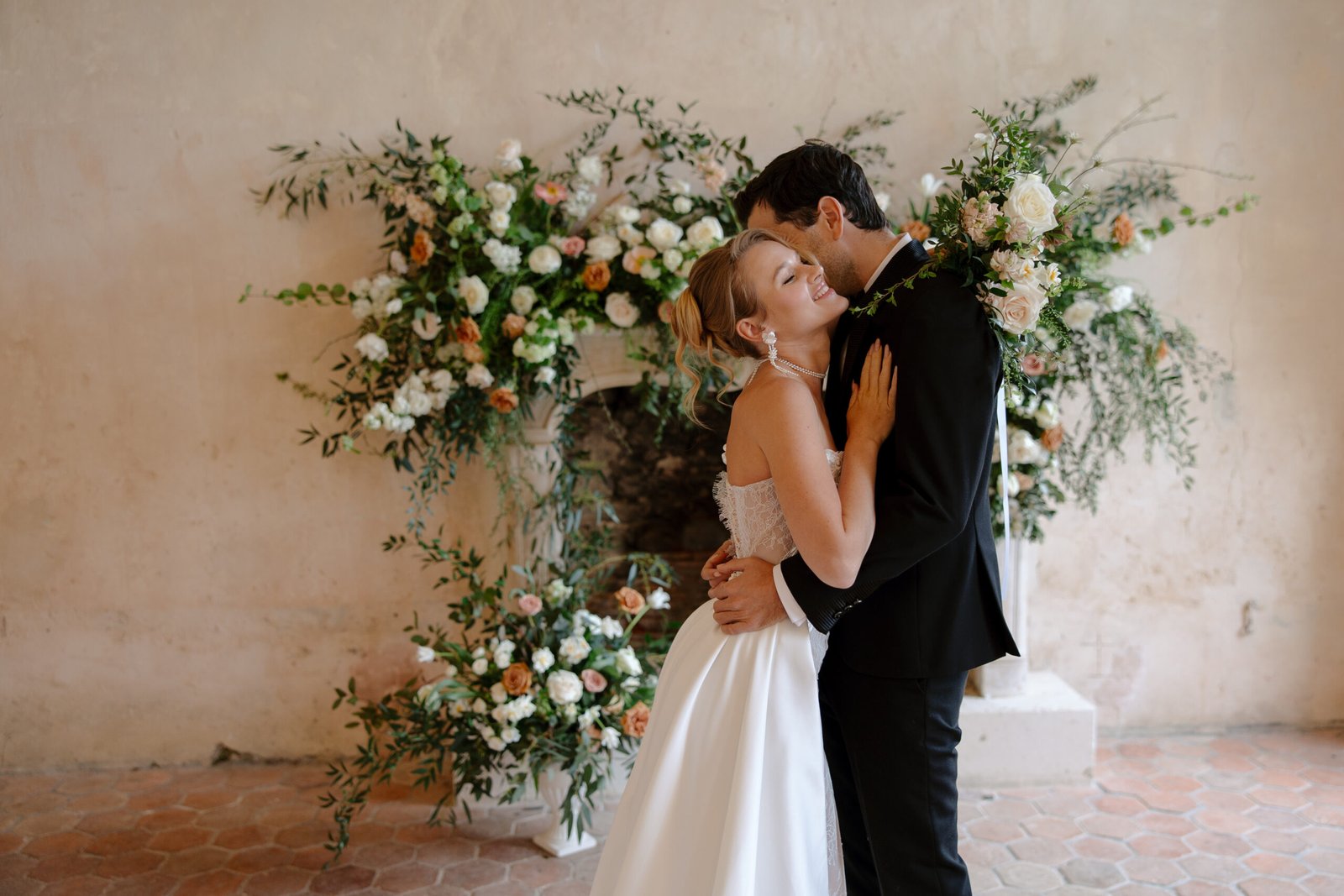 A bride and groom embrace in front of a floral arrangement, both smiling, with the bride in a white dress and the groom in a black suit.