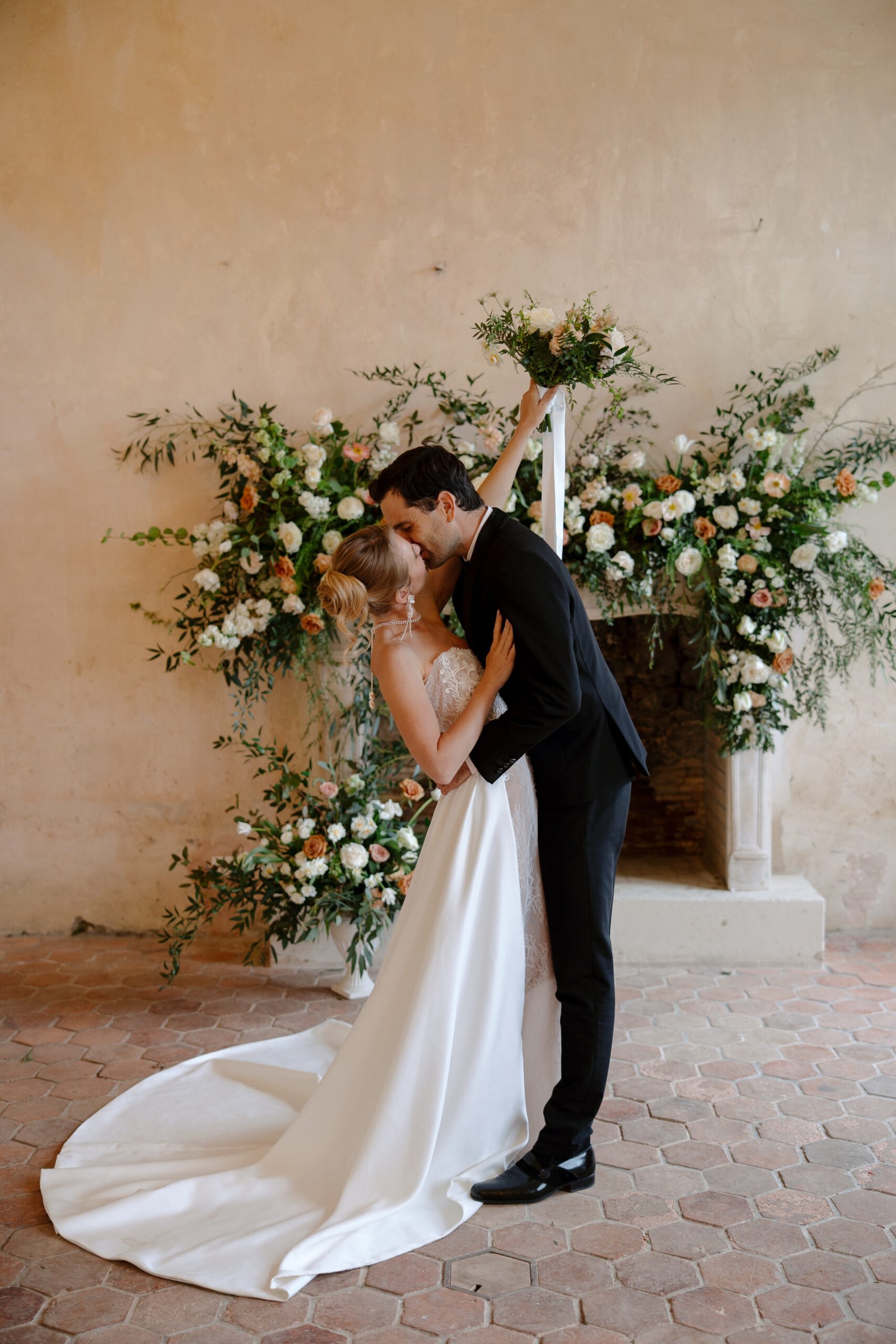 A bride and groom share a kiss in front of a flower-adorned backdrop; the bride holds a bouquet aloft while wearing a white gown, and the groom is in a black suit. Chateau de Courtomer wedding photographer.
