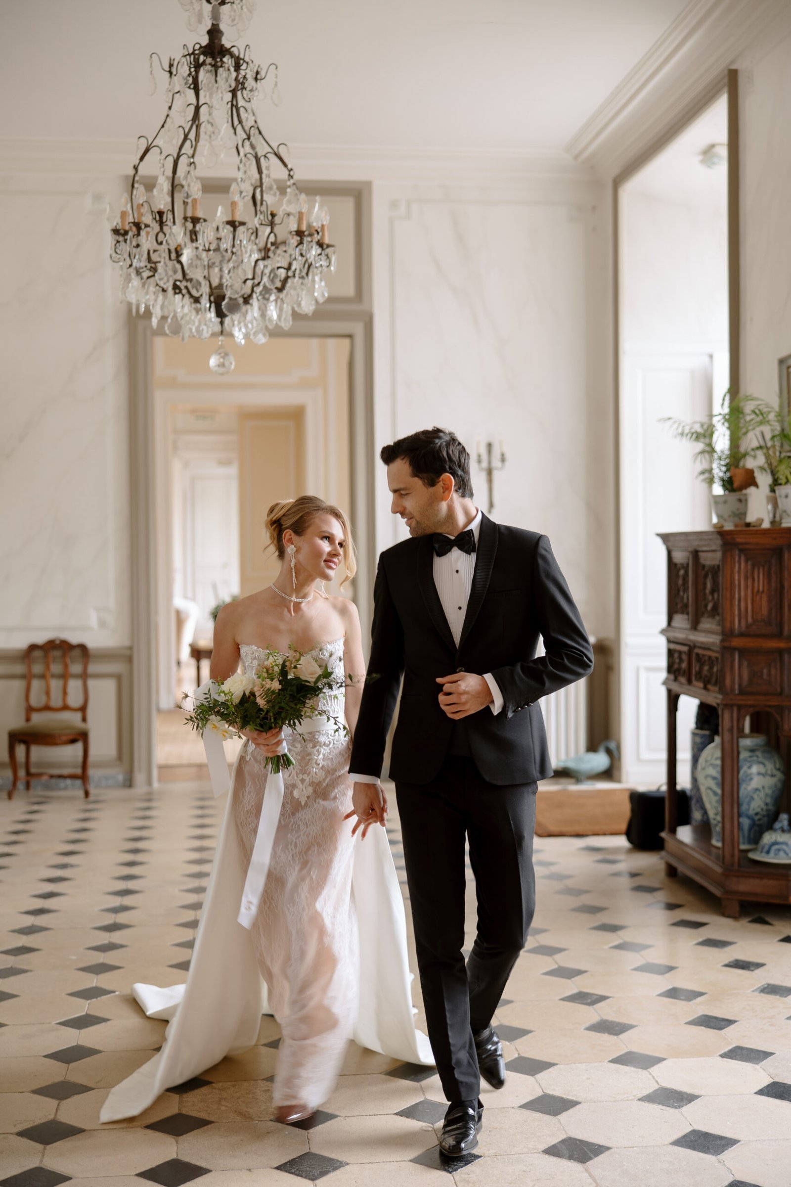A bride holding a bouquet and a groom in a tuxedo walk together in an elegant, well-lit room with checkered flooring and a chandelier.