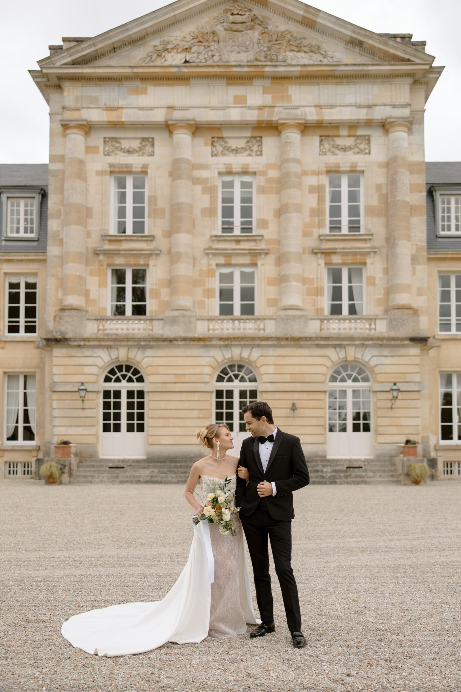 A bride and groom stand together outside a grand, classical-style building, looking at each other and holding a bouquet. Captured by France wedding photographer.