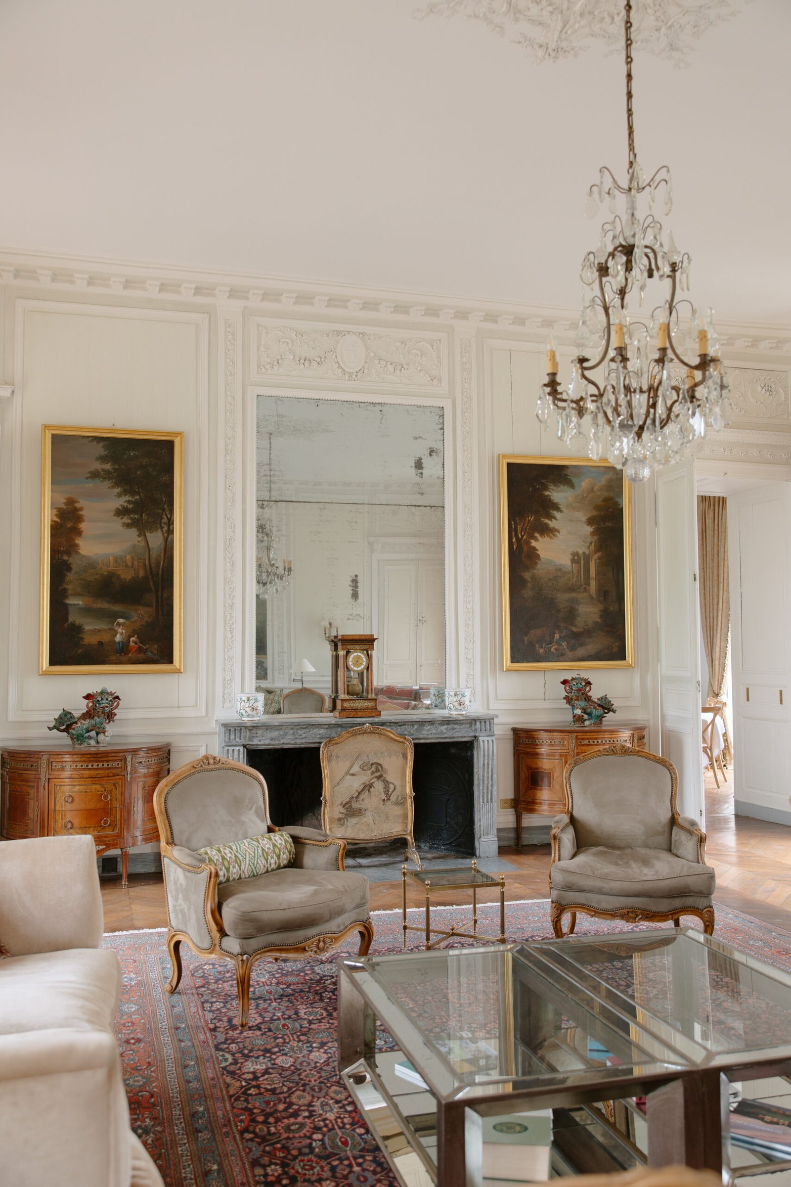 Elegant living room with ornate white walls, a chandelier, two armchairs, a large mirror above a fireplace, classic paintings, and vintage wooden furniture on a patterned rug.