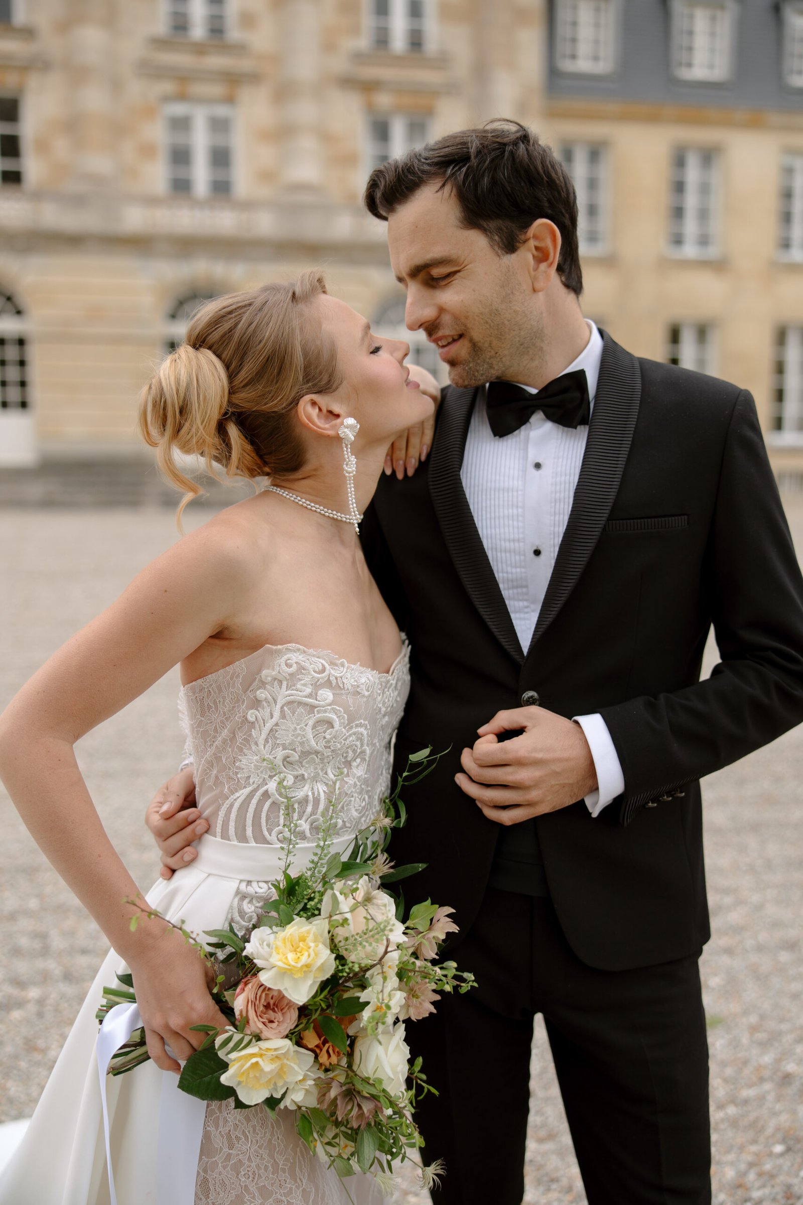 A bride in a white wedding dress and a groom in a black tuxedo stand close together outdoors, holding a bouquet and looking at each other. Chateau de Courtomer wedding photographer.