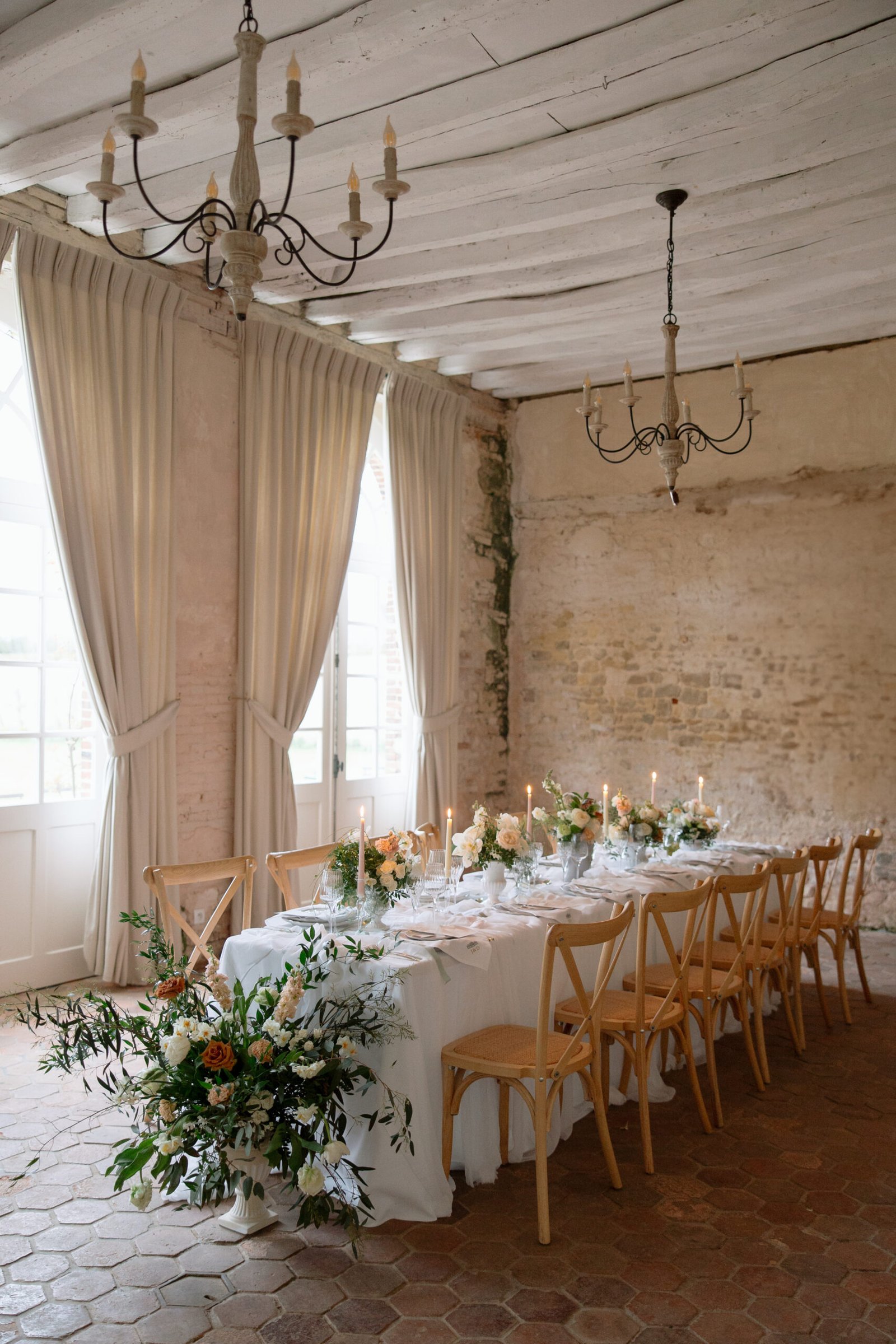 A long dining table set with white linens, candles, and floral arrangements is surrounded by wooden chairs in a rustic room with high ceilings, tall windows, and exposed brick walls.