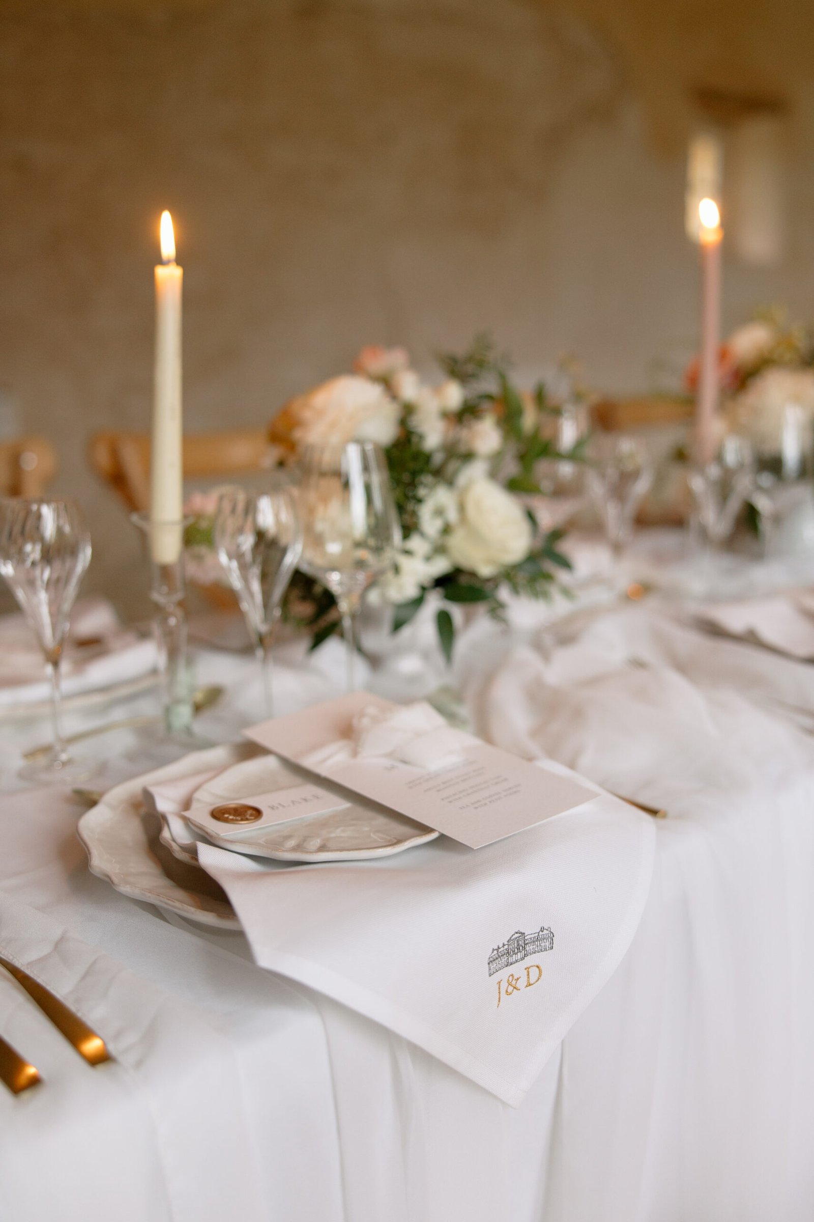 Elegant table setting with white linens, floral centerpiece, gold cutlery, wine glasses, lit candles, and monogrammed napkins marked J & D. Menus are placed on the plates.
