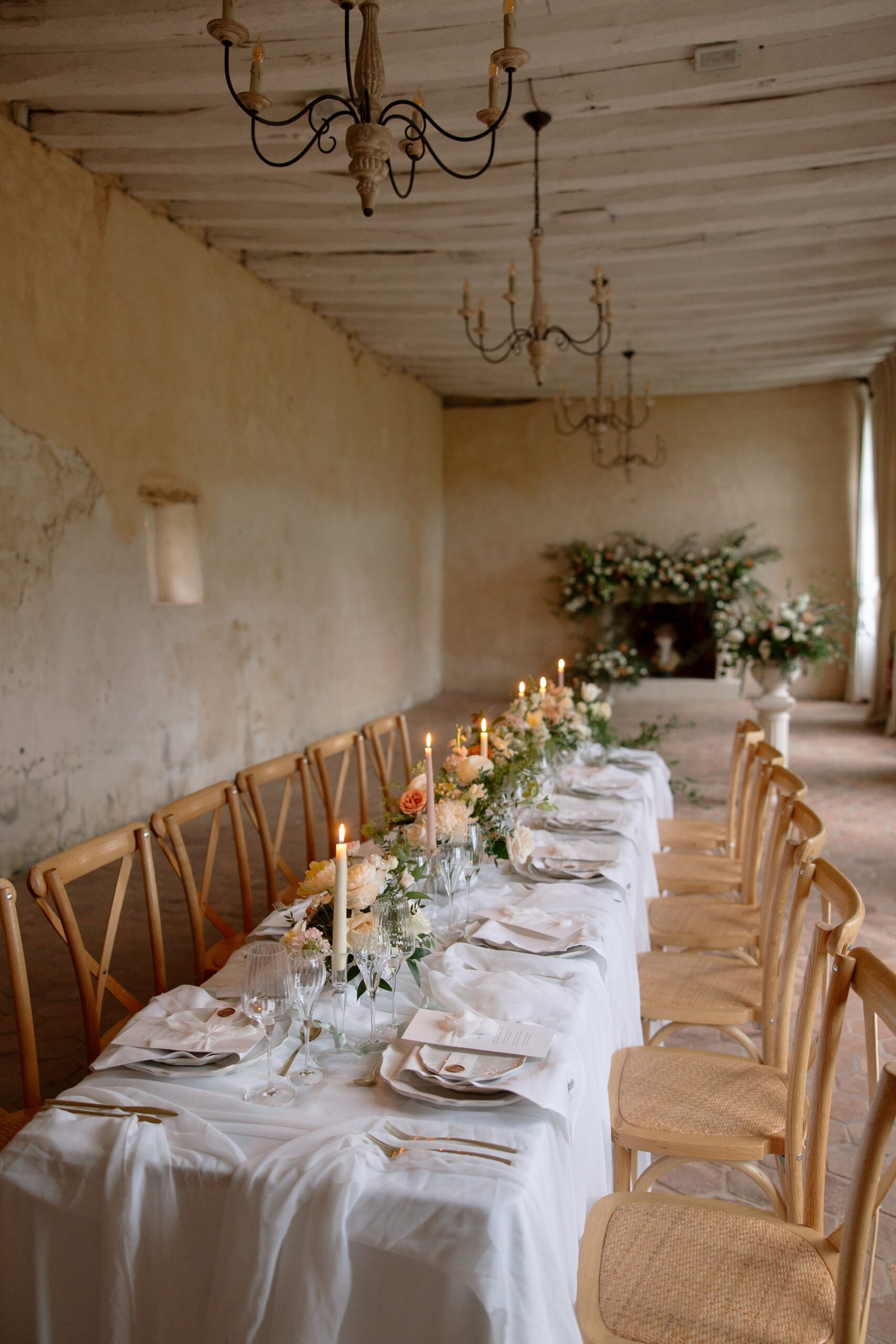 A long dining table with a white tablecloth, set with plates, glassware, candles, and floral centerpieces, stands in a rustic room with wooden chairs and chandeliers.