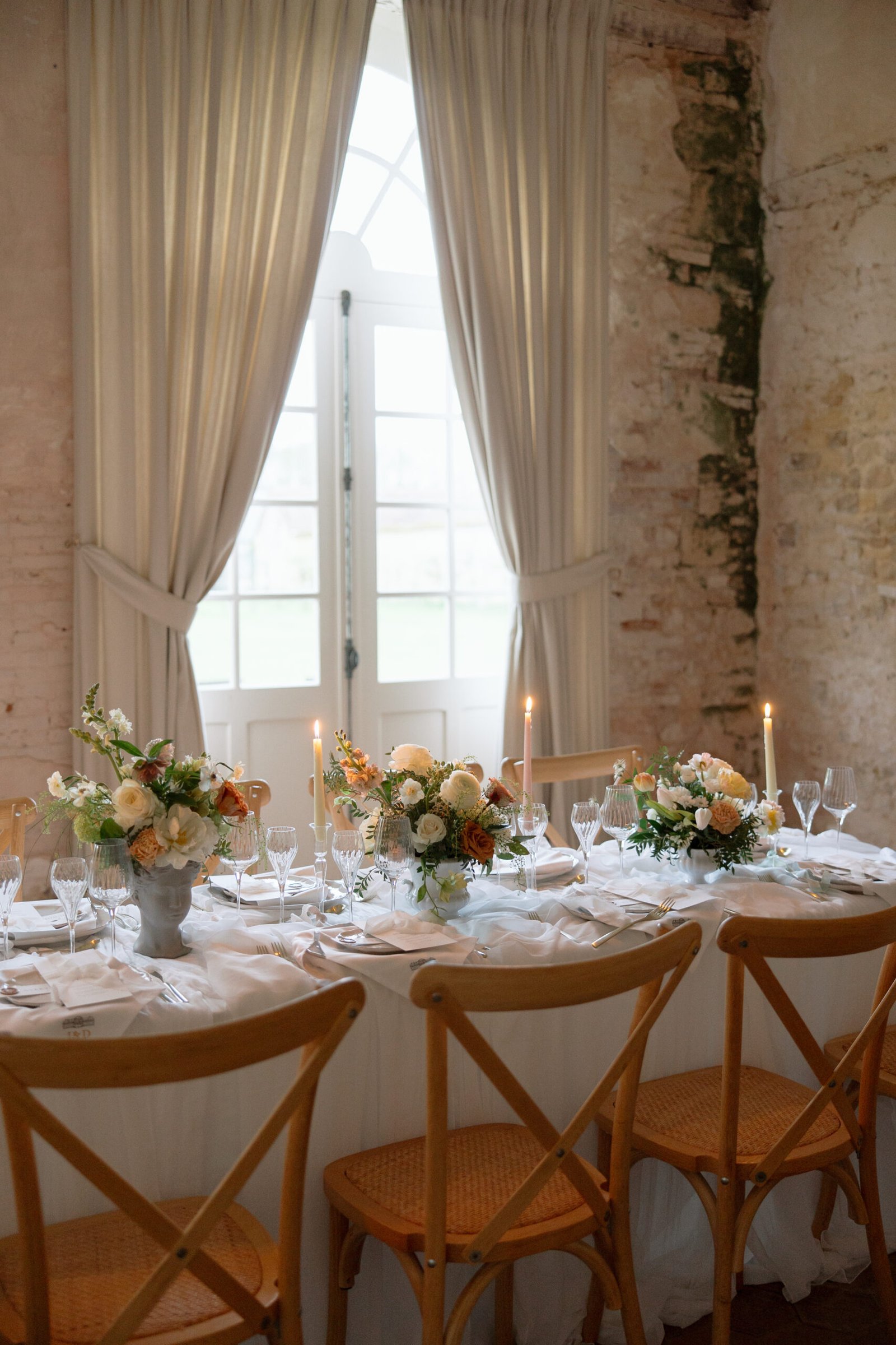 A table set for a formal event with floral centerpieces, candles, glassware, and wooden chairs in front of a window with sheer curtains and textured walls.