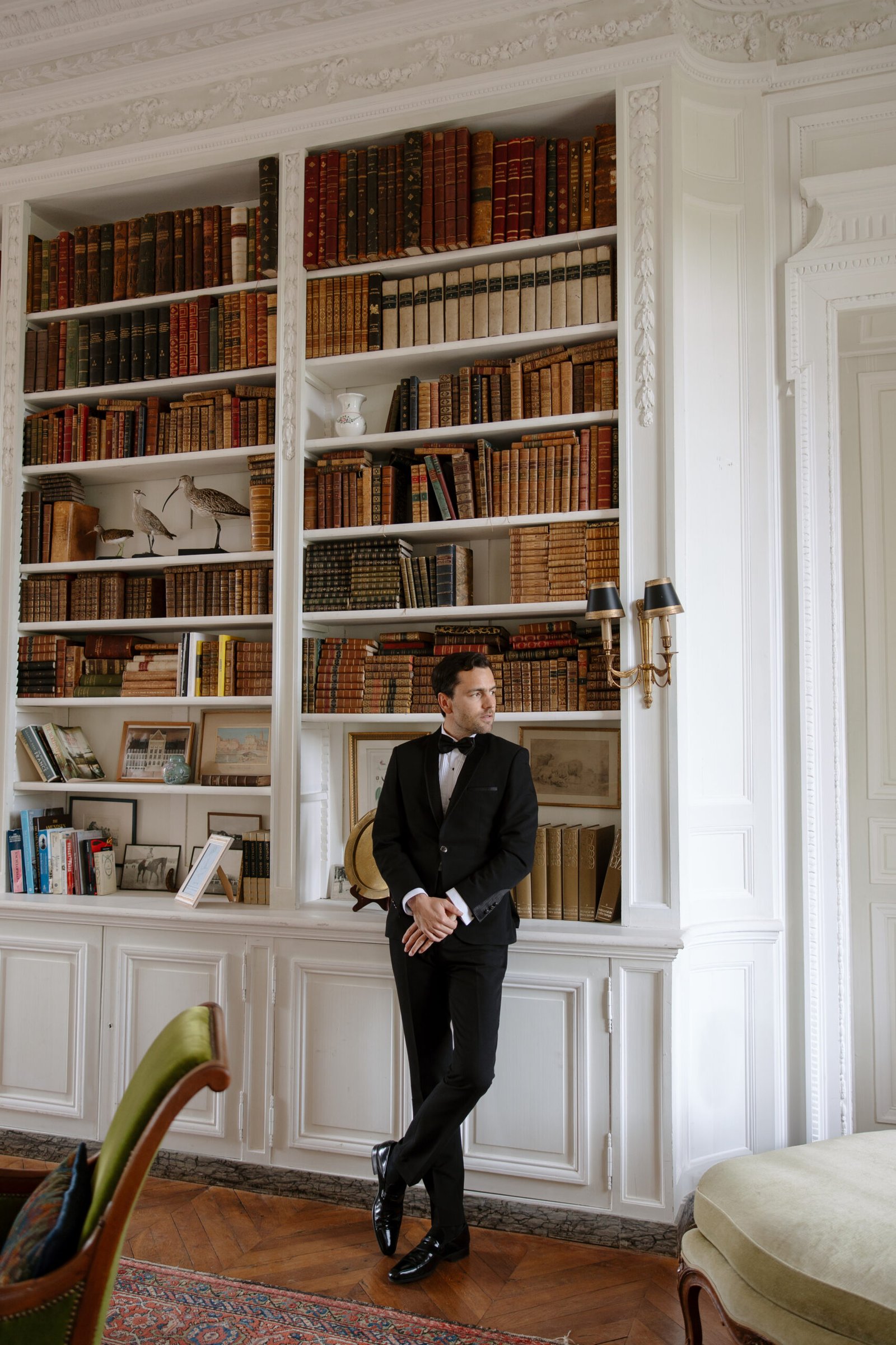A man in a black tuxedo stands in front of a white built-in bookshelf filled with books and decorative items in a formal room.