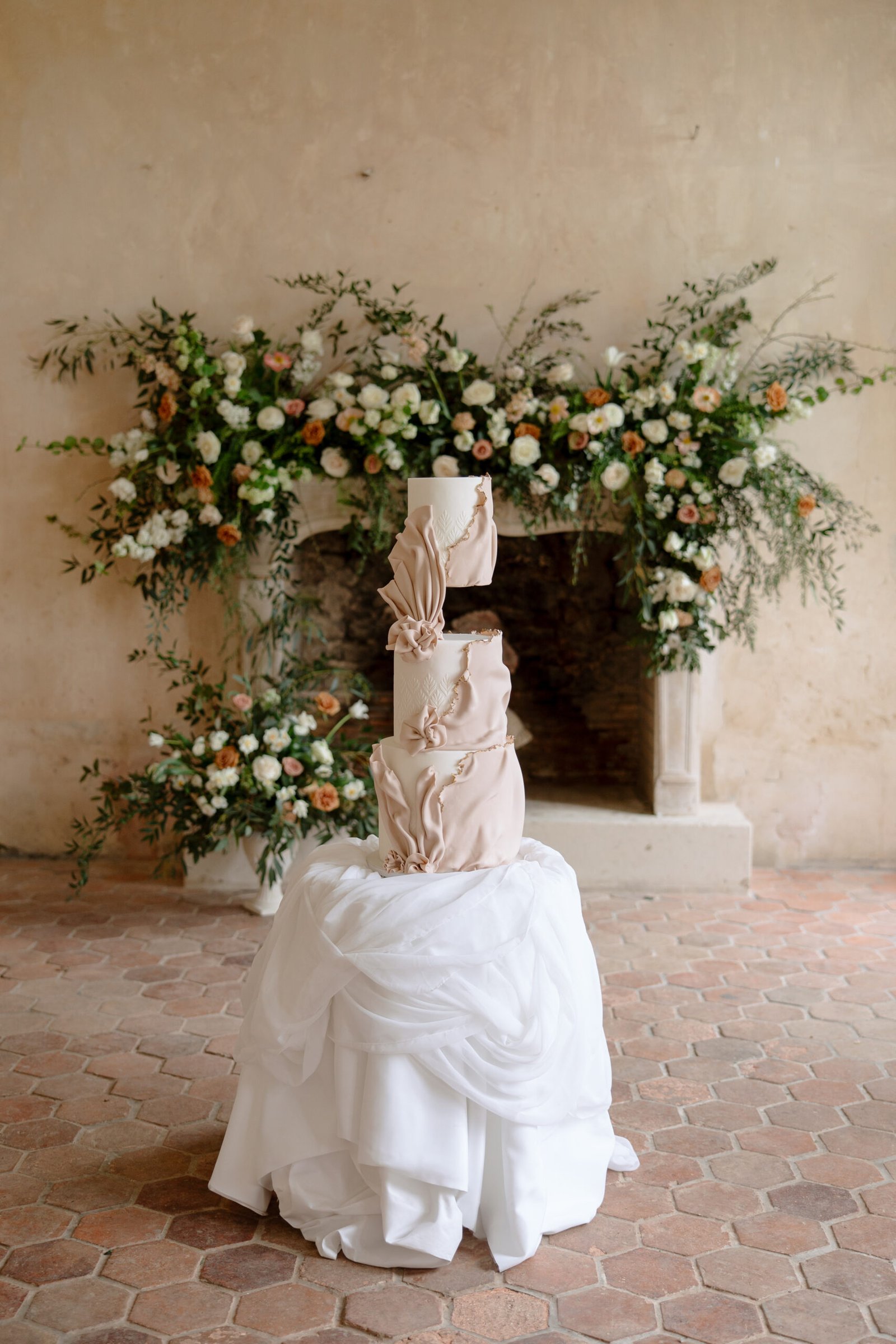 Three-tier beige wedding cake with draped fondant and flower accents, displayed on a round table with a white cloth, set in front of a floral arrangement and stone fireplace.