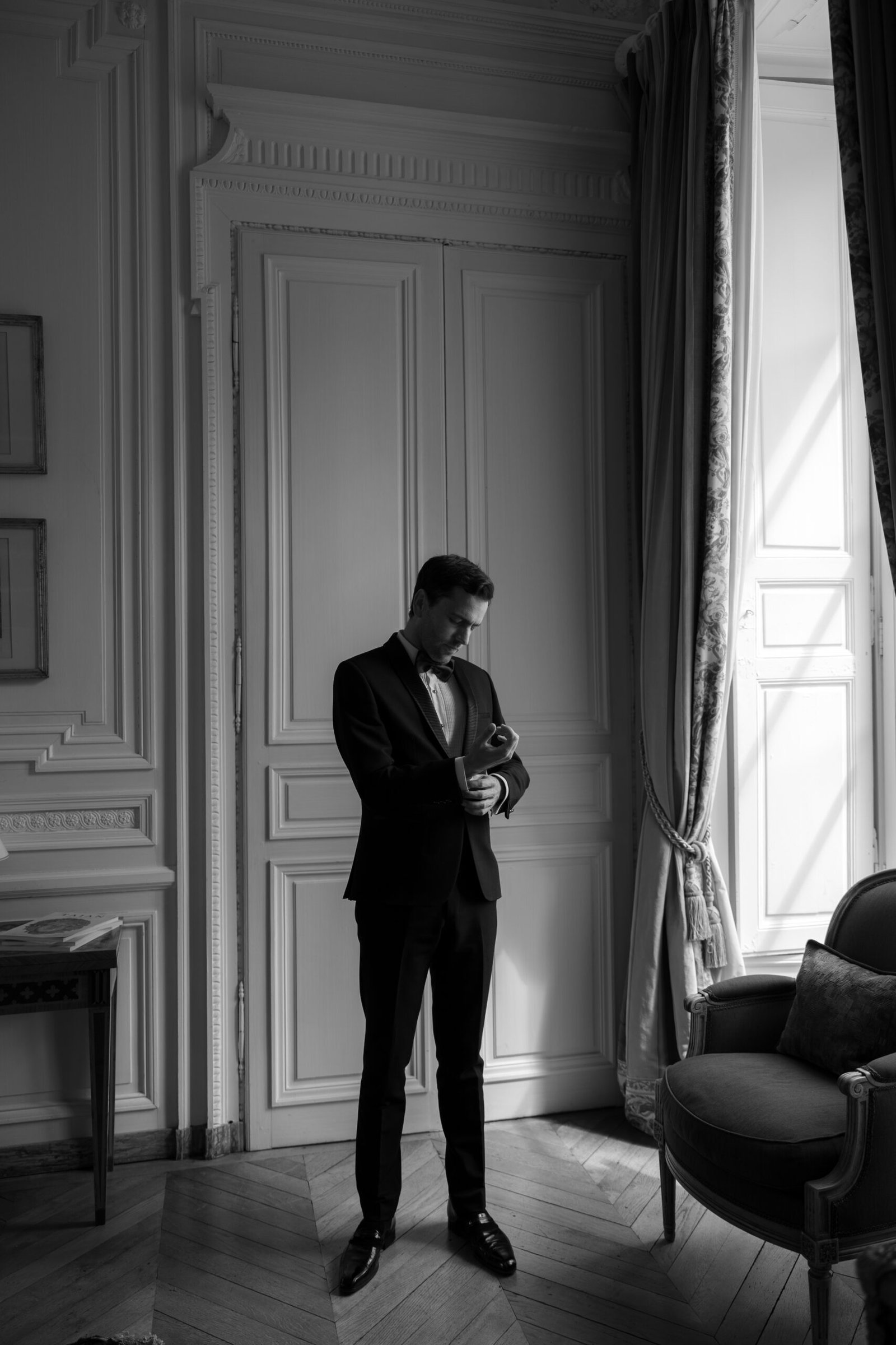 A man in a suit stands by a window in an elegant room, adjusting his watch and looking down, with light streaming in from the side.