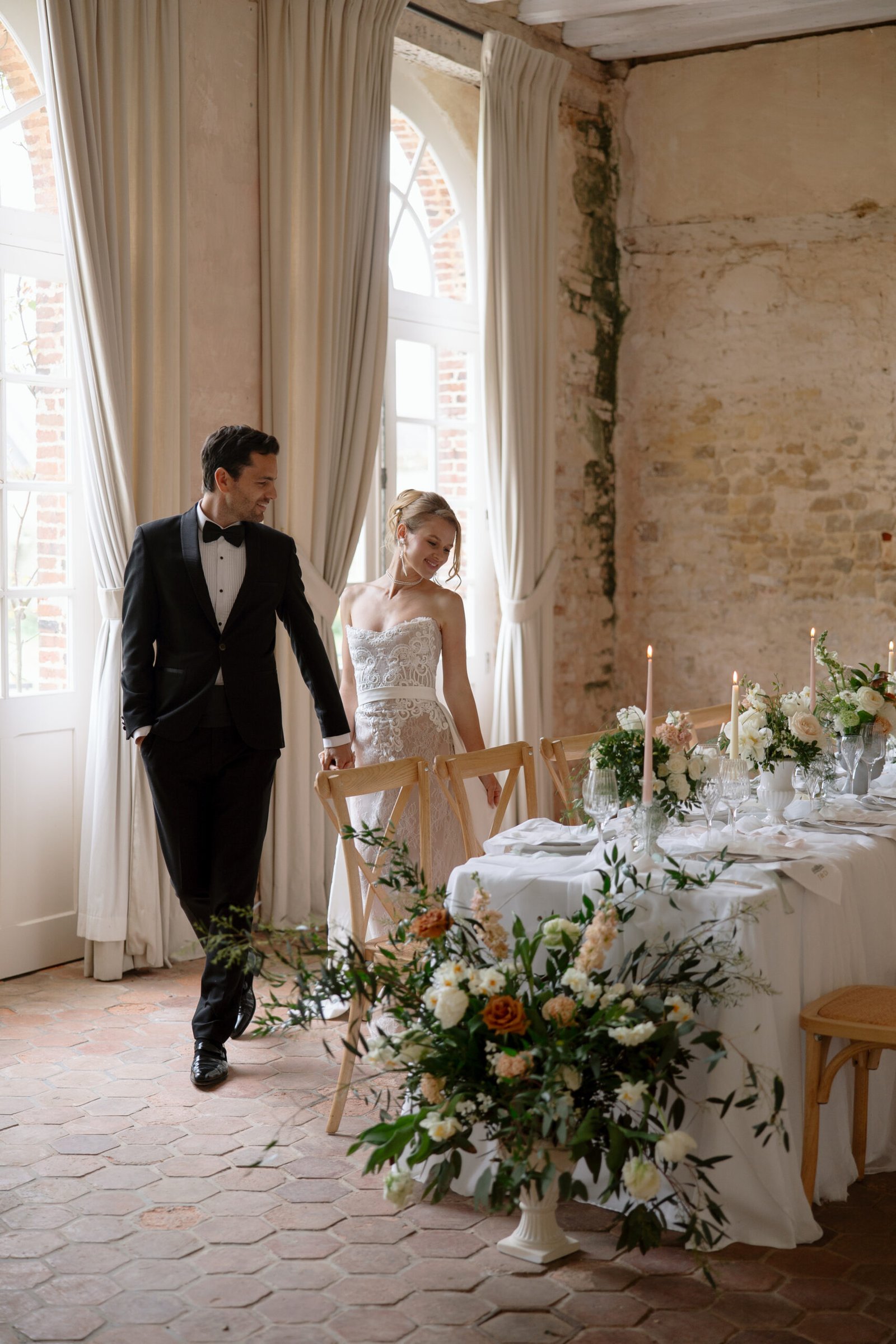 A bride and groom in formal attire stand by an elegantly set dining table with floral arrangements in a rustic, light-filled room.