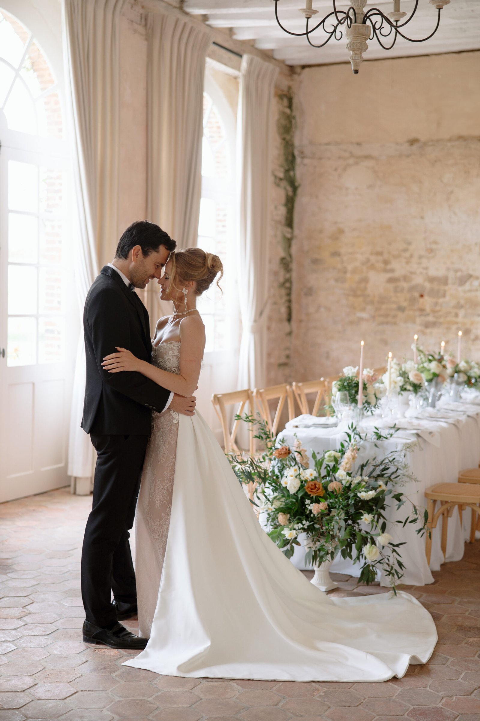 A bride and groom stand close together in an elegant, light-filled room with a decorated table set for a wedding reception.
