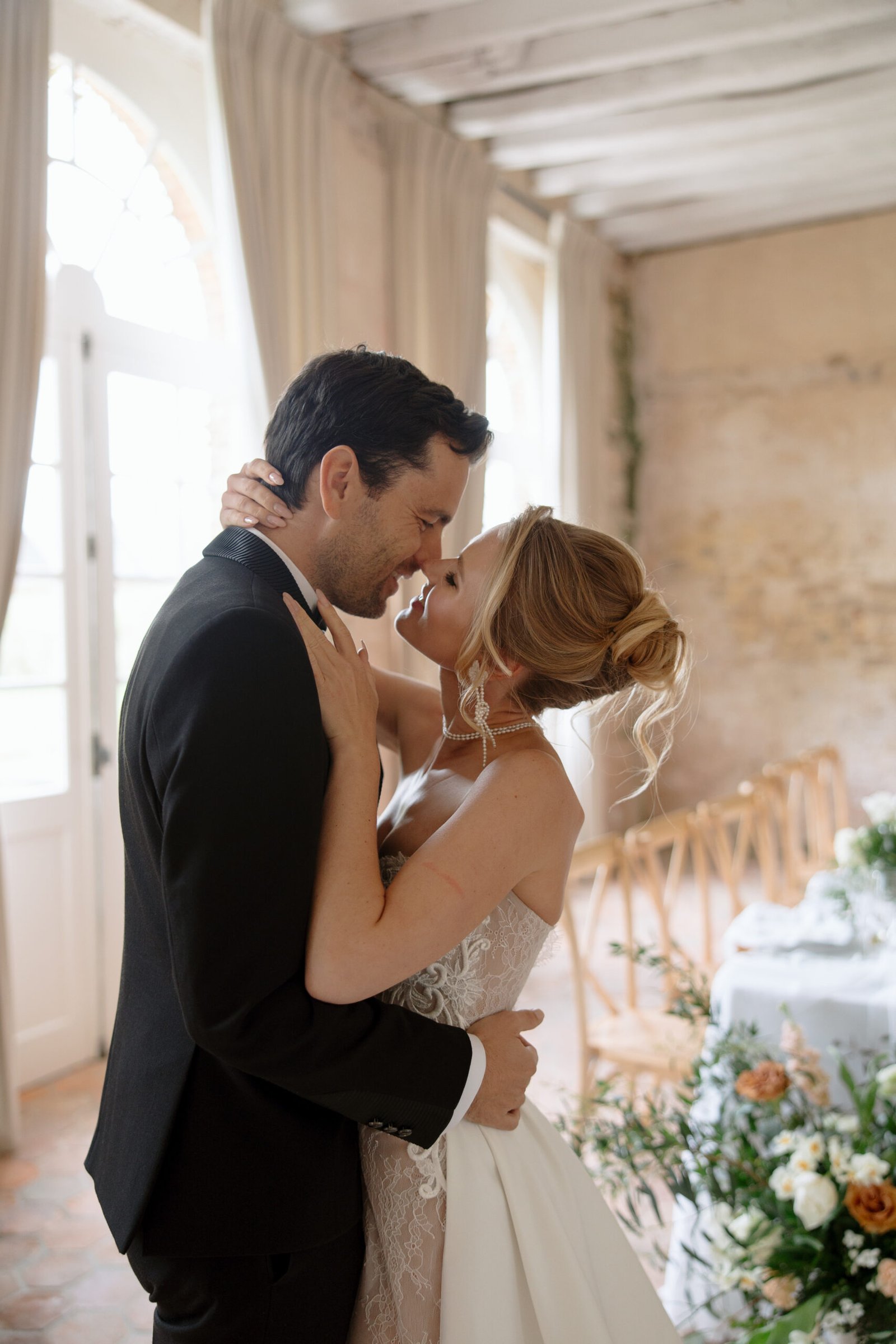 A bride and groom embrace and smile at each other in a softly lit, elegant room decorated for a wedding.