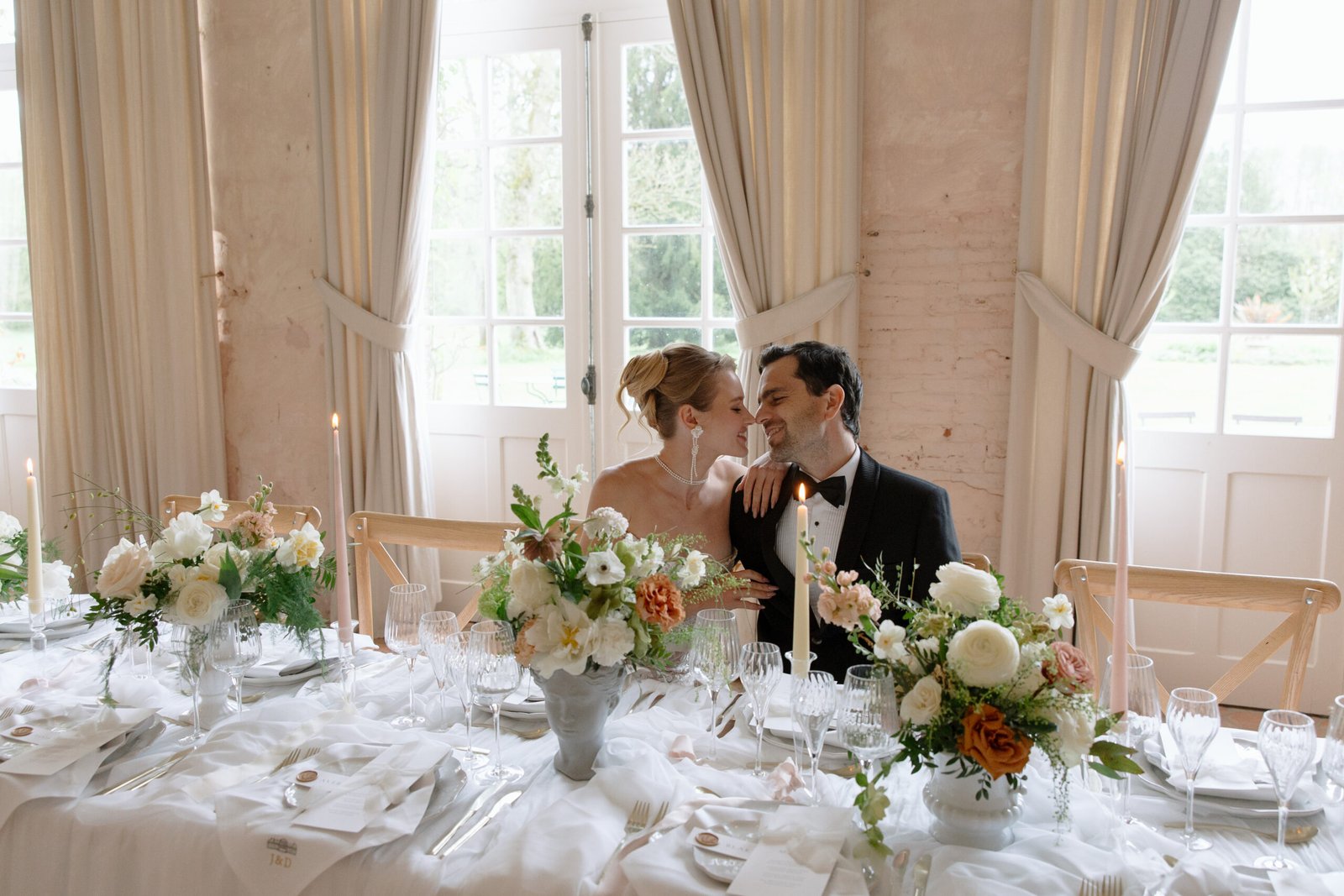 A bride and groom sit closely together at a decorated wedding reception table with floral arrangements and candles in front of large windows.