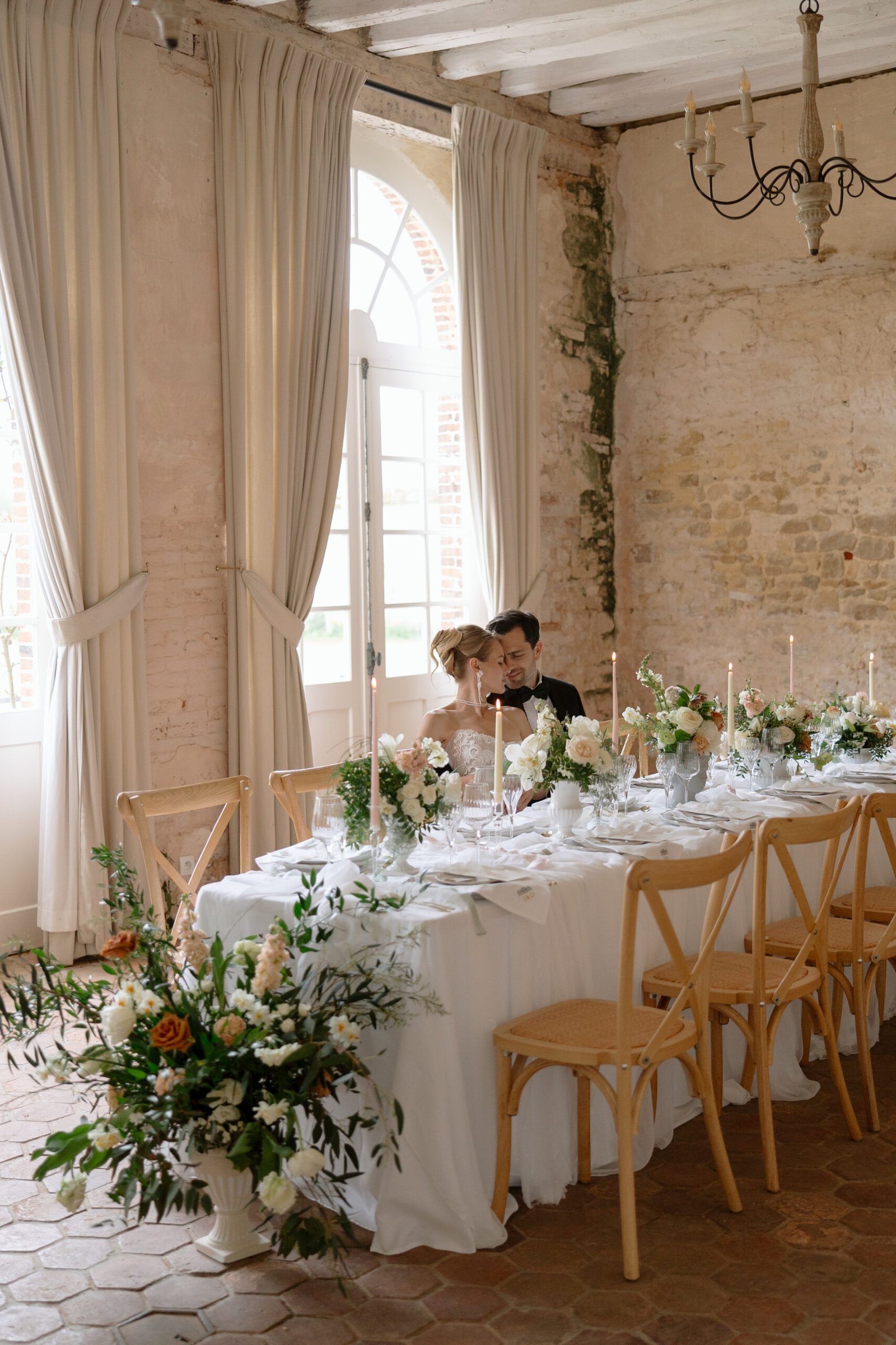 A bride and groom sit together at a decorated table set for a wedding reception in a rustic room with large windows and cream curtains. Chateau de Courtomer wedding photographer.