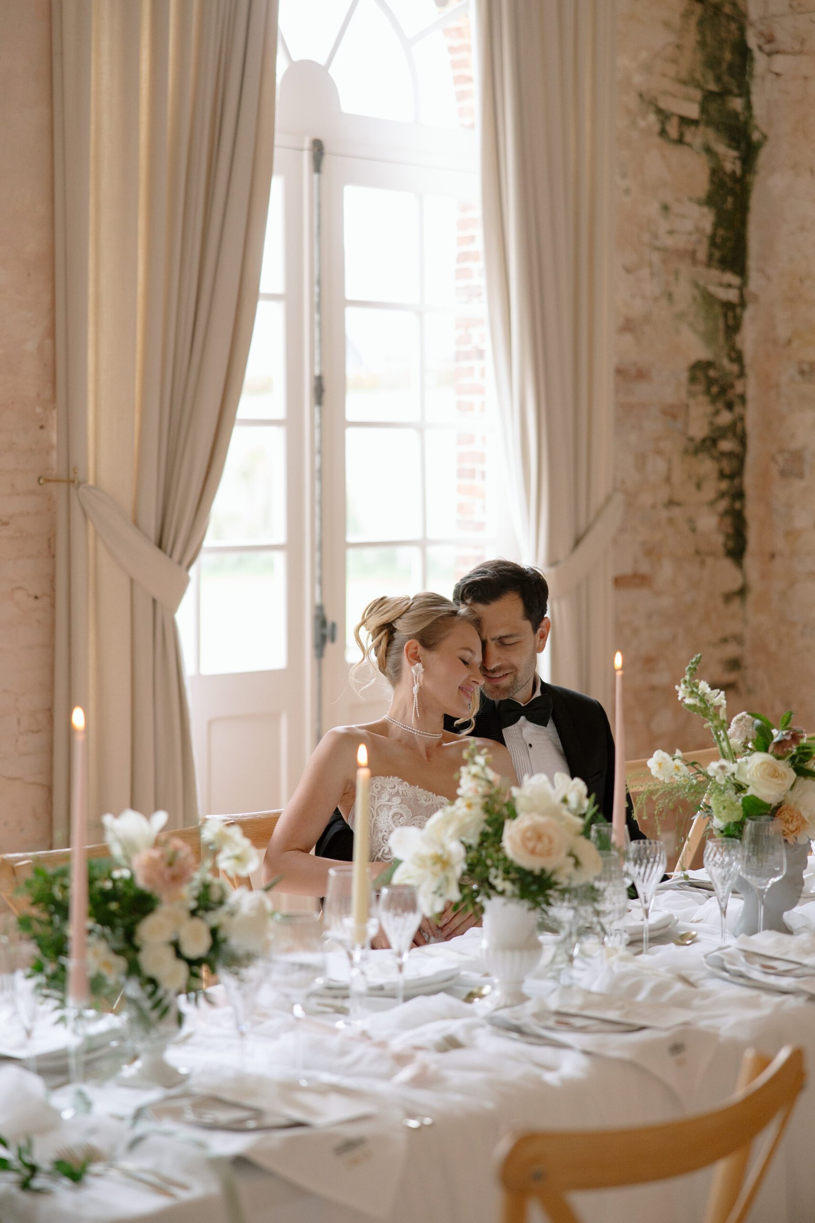 A couple dressed in formal attire sits closely together at an elegantly decorated table with flowers, candles, and glassware in a softly lit room with large windows and brick walls.