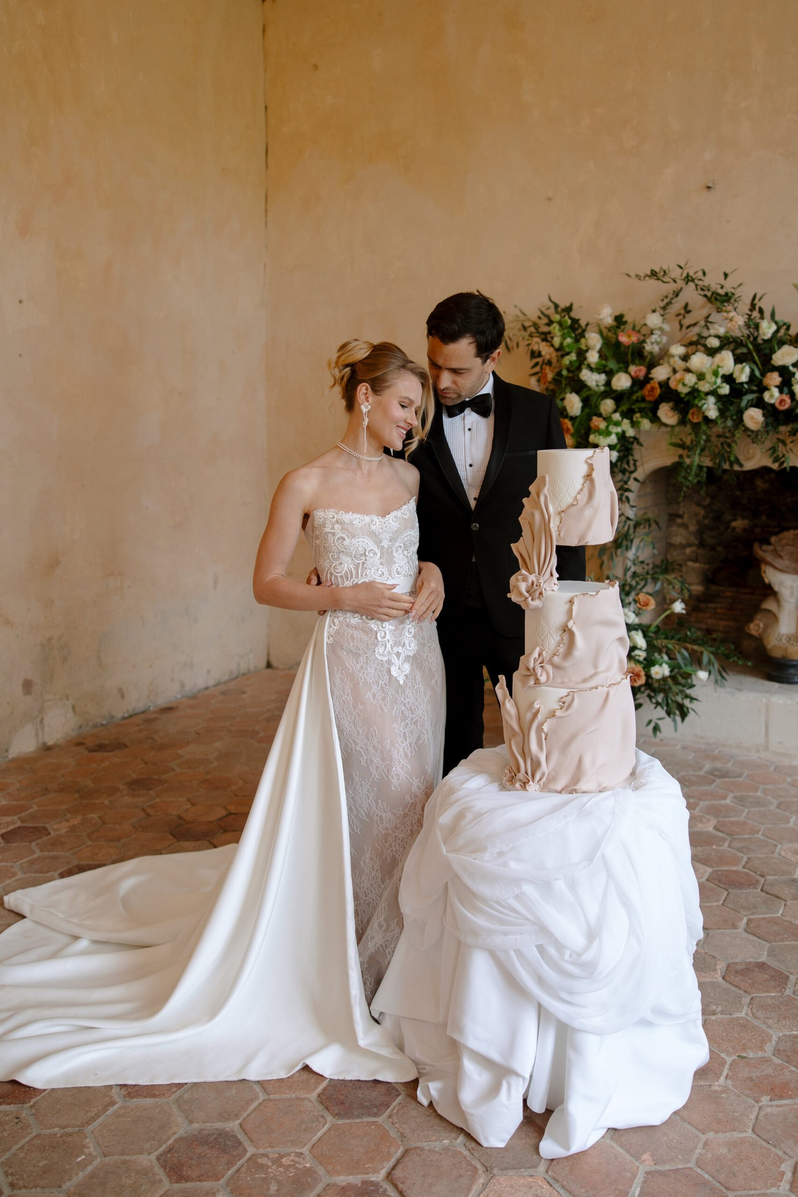 A bride and groom in formal attire stand beside a multi-tiered wedding cake in an elegant indoor setting with floral arrangements.