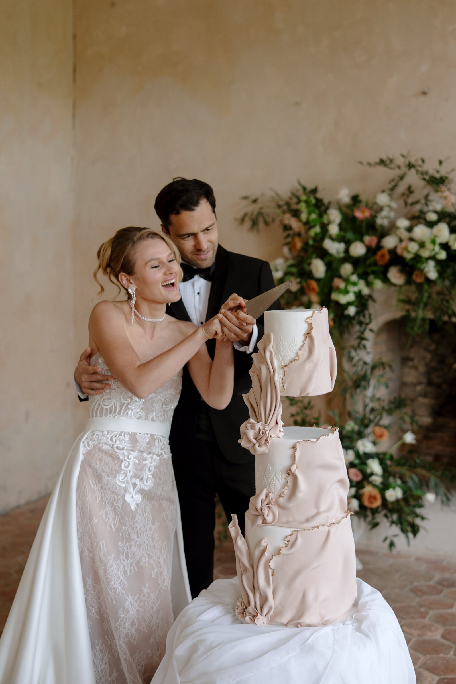 A bride and groom, dressed in formal wedding attire, smile as they cut a multi-tiered pink wedding cake together.