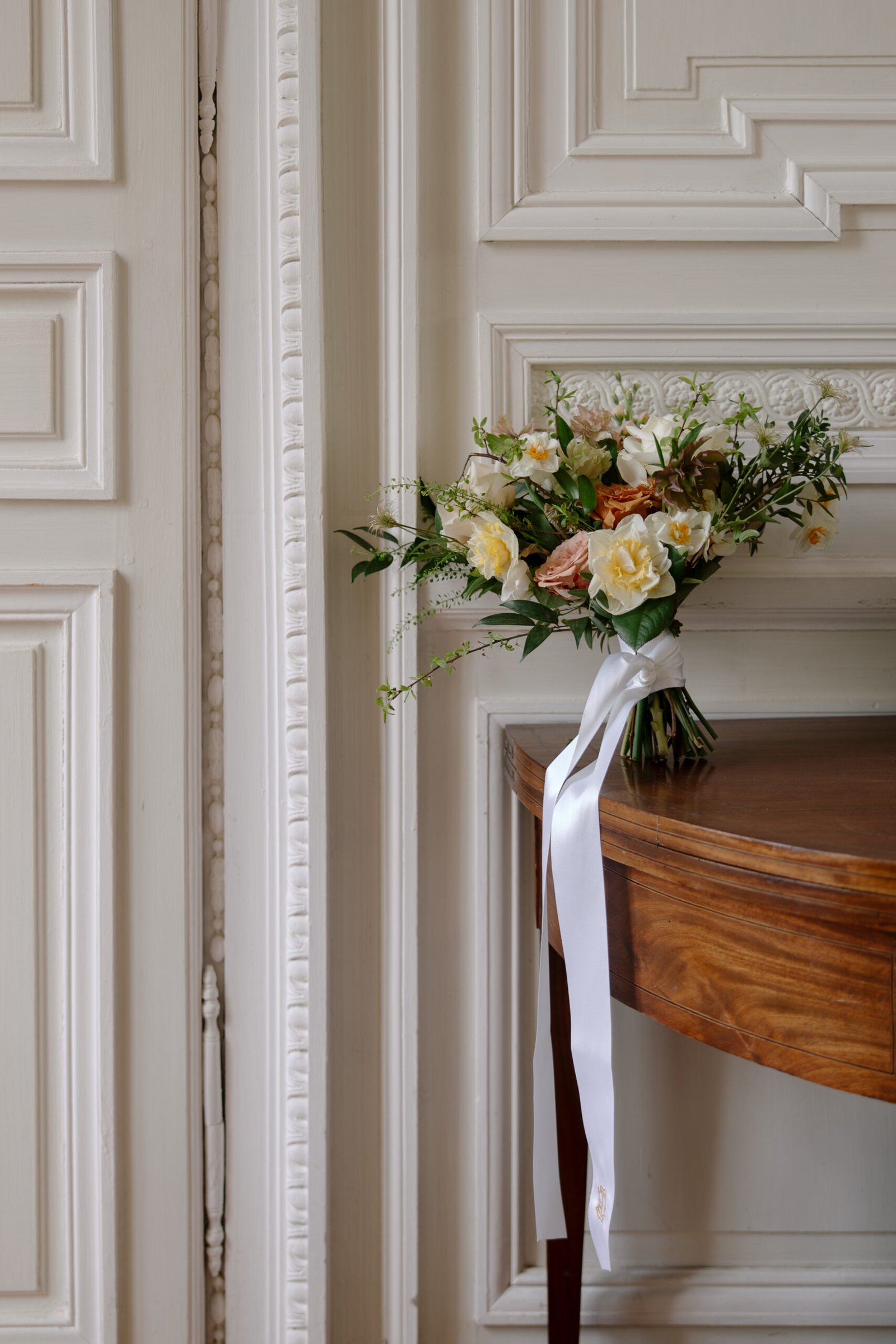 A bouquet of white and pale yellow flowers with greenery and a long white ribbon rests on the edge of a wooden table beside an ornate white paneled wall.