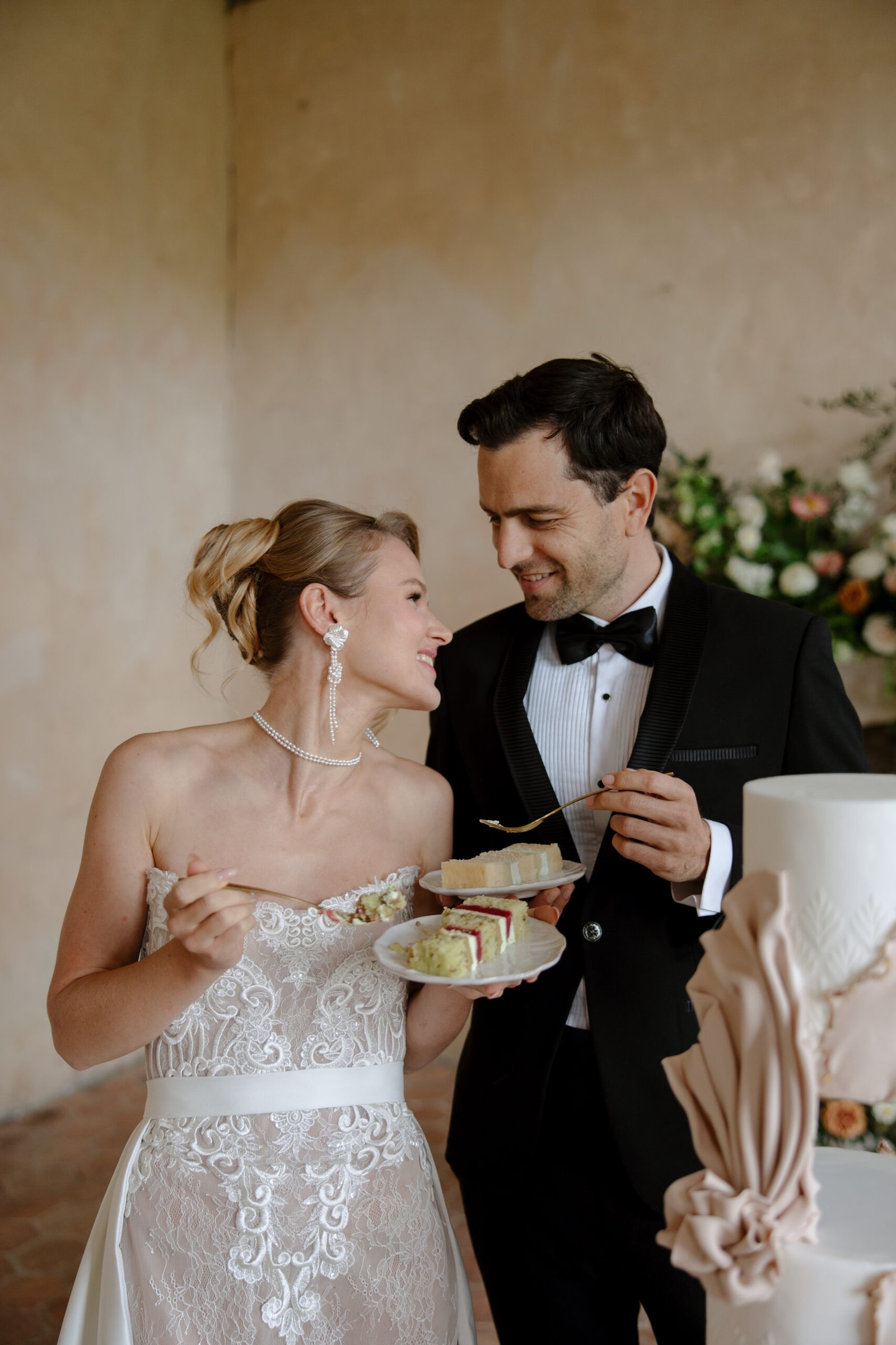 A bride and groom in formal attire stand together, smiling and holding plates of wedding cake, with a large decorated cake in the foreground.