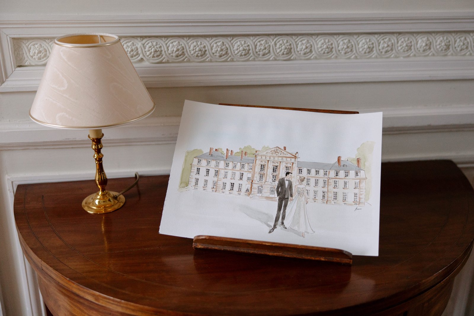 A watercolor painting of a couple in wedding attire stands on a wooden table next to a brass lamp, with ornate wall molding in the background.