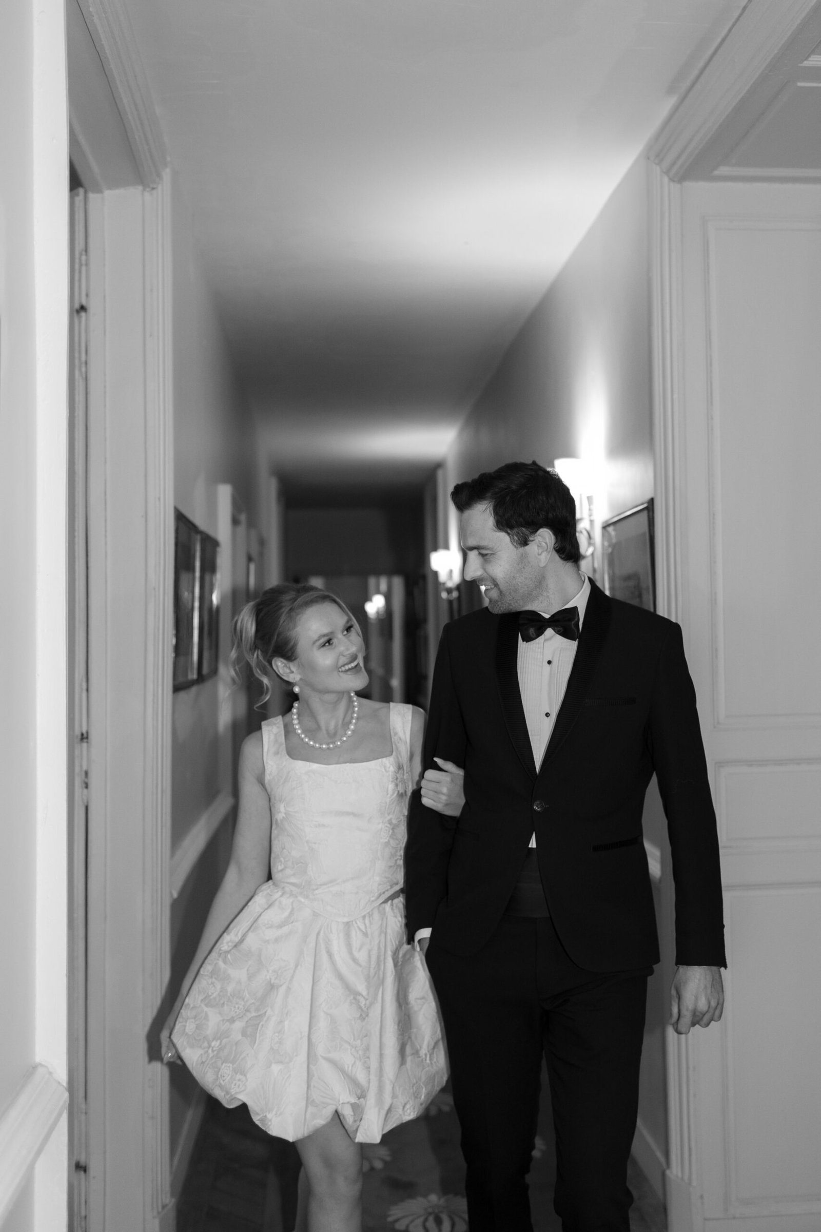 A woman in a short, white dress and a man in a black suit with a bow tie walk arm in arm down a hallway, looking at each other and smiling. Chateau de Courtomer wedding photographer.