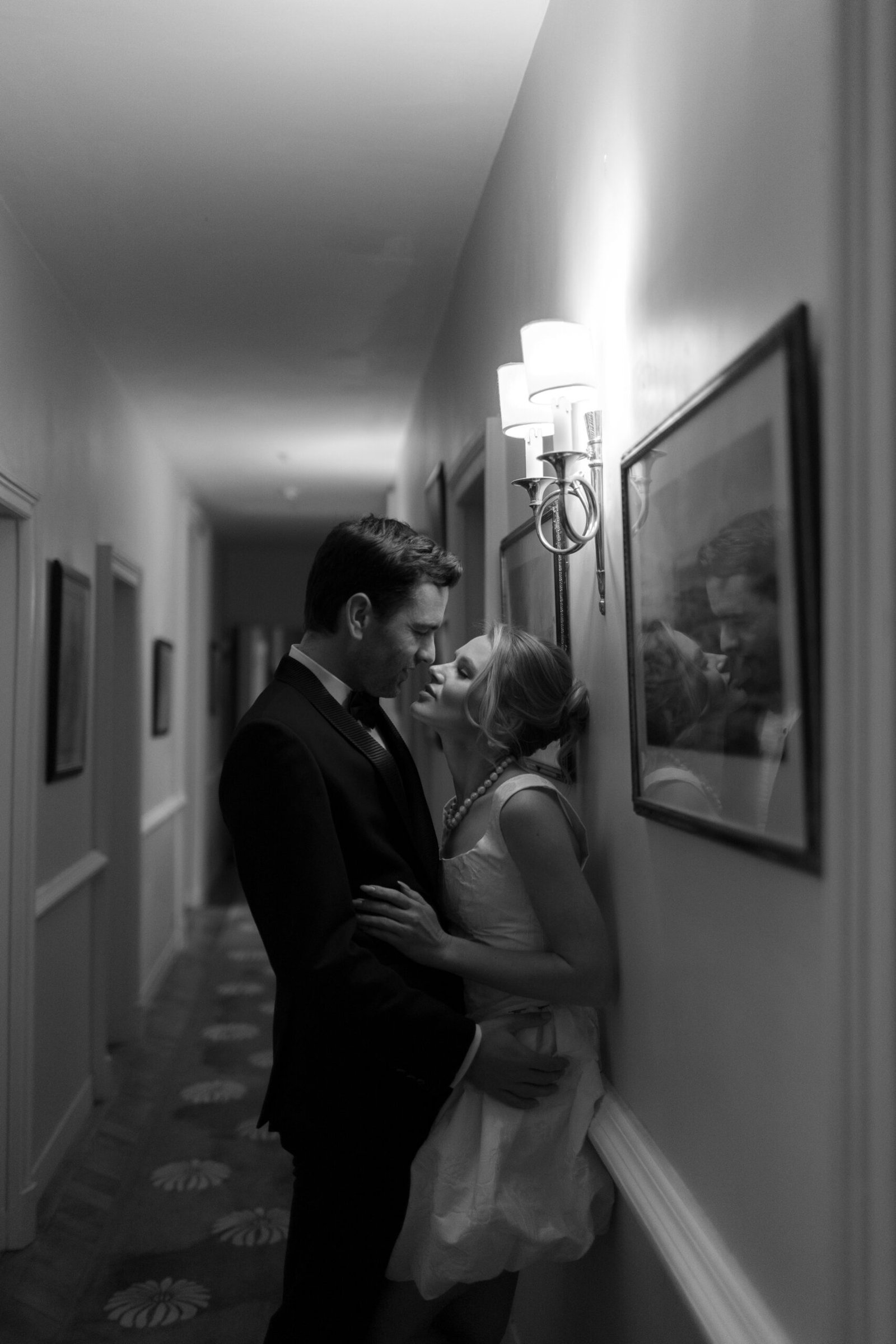 A man in a suit and a woman in a dress stand closely together in a dimly lit hallway, sharing an intimate moment near a wall sconce and framed pictures.