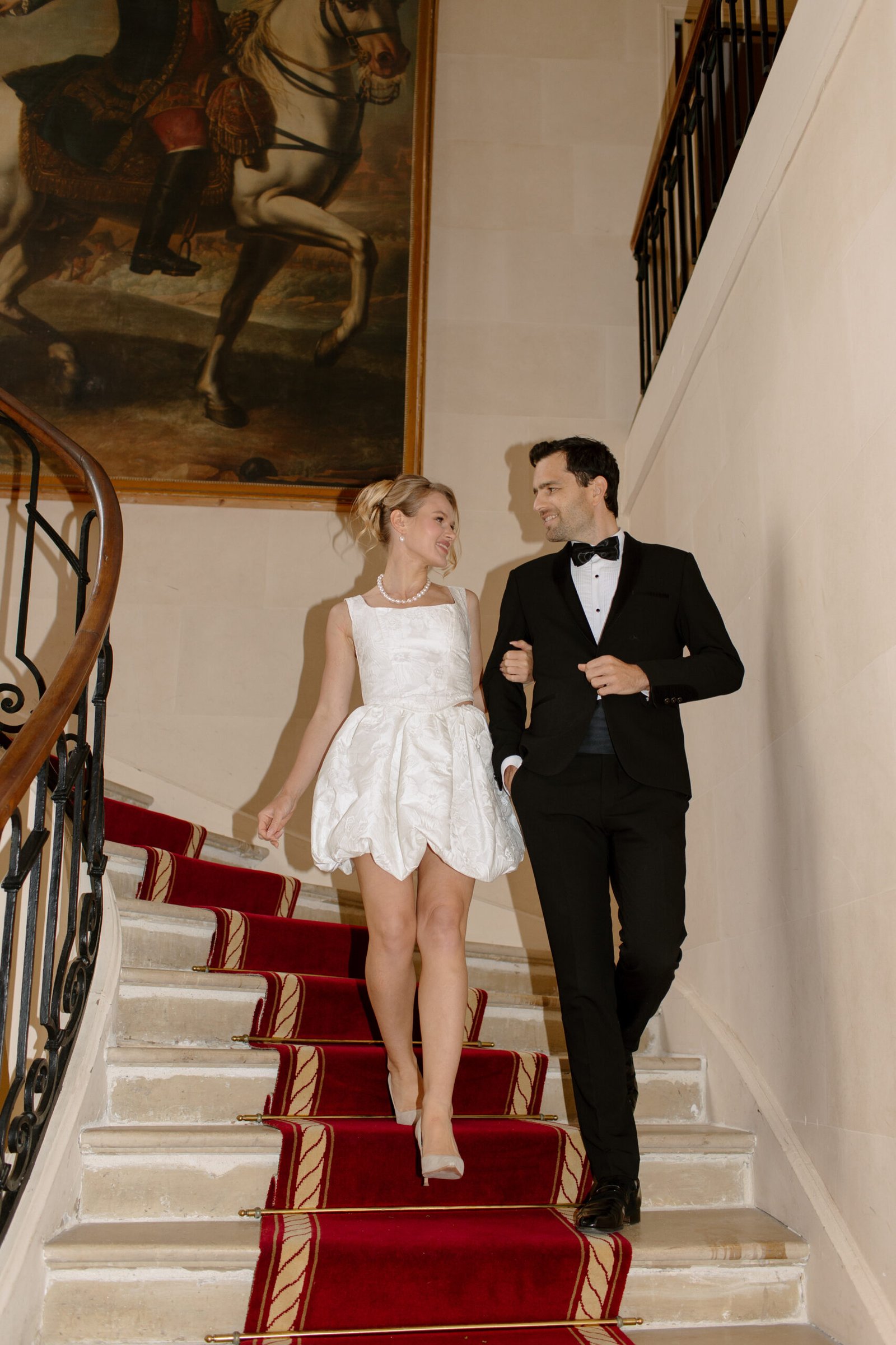 A woman in a short white dress and a man in a black tuxedo walk down a red-carpeted staircase, smiling at each other, with a large painting on the wall behind them. Chateau de Courtomer wedding photographer.