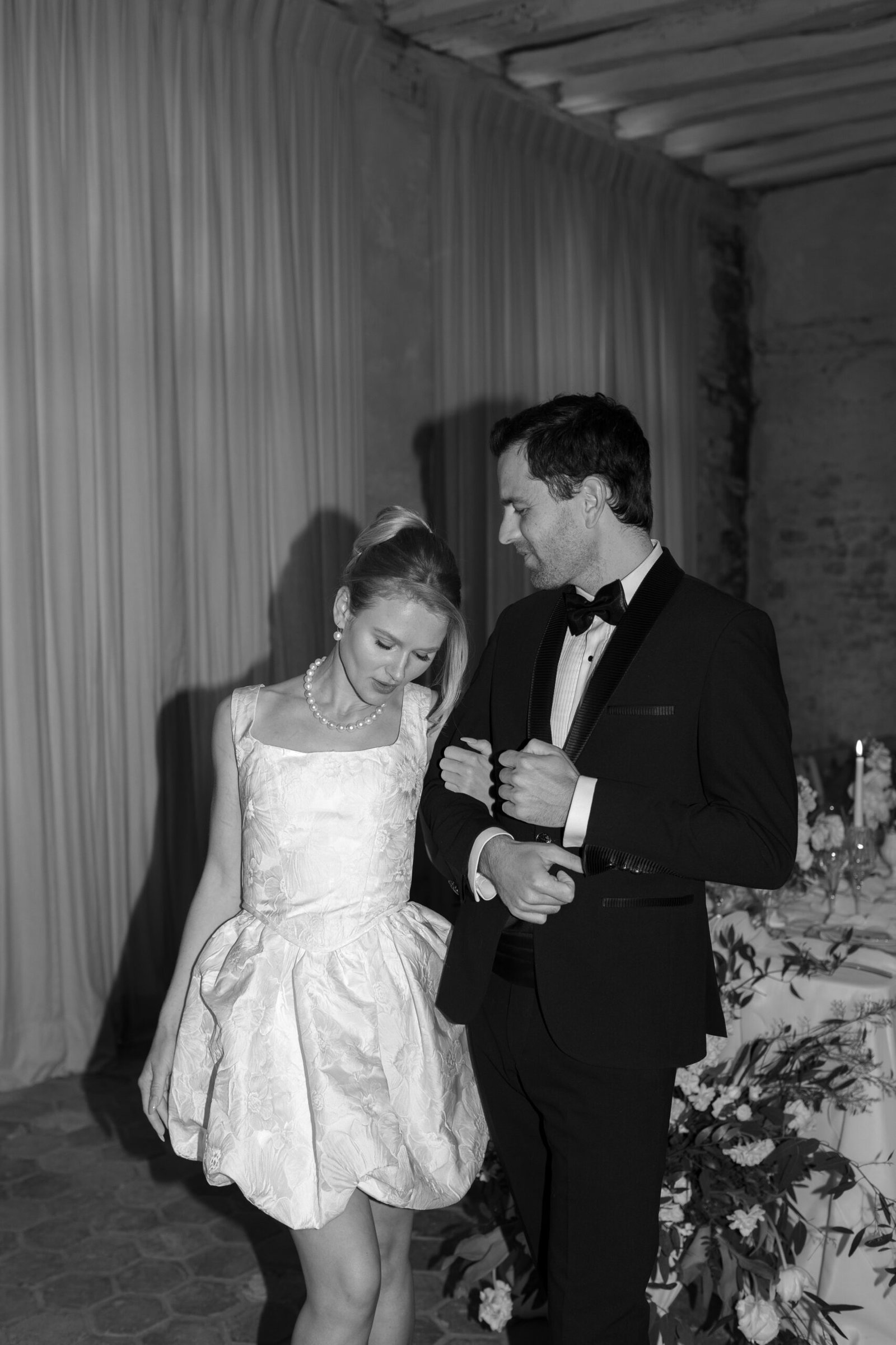 A woman in a short white dress stands beside a man in a black tuxedo; they are indoors near a decorated table with flowers and candles.