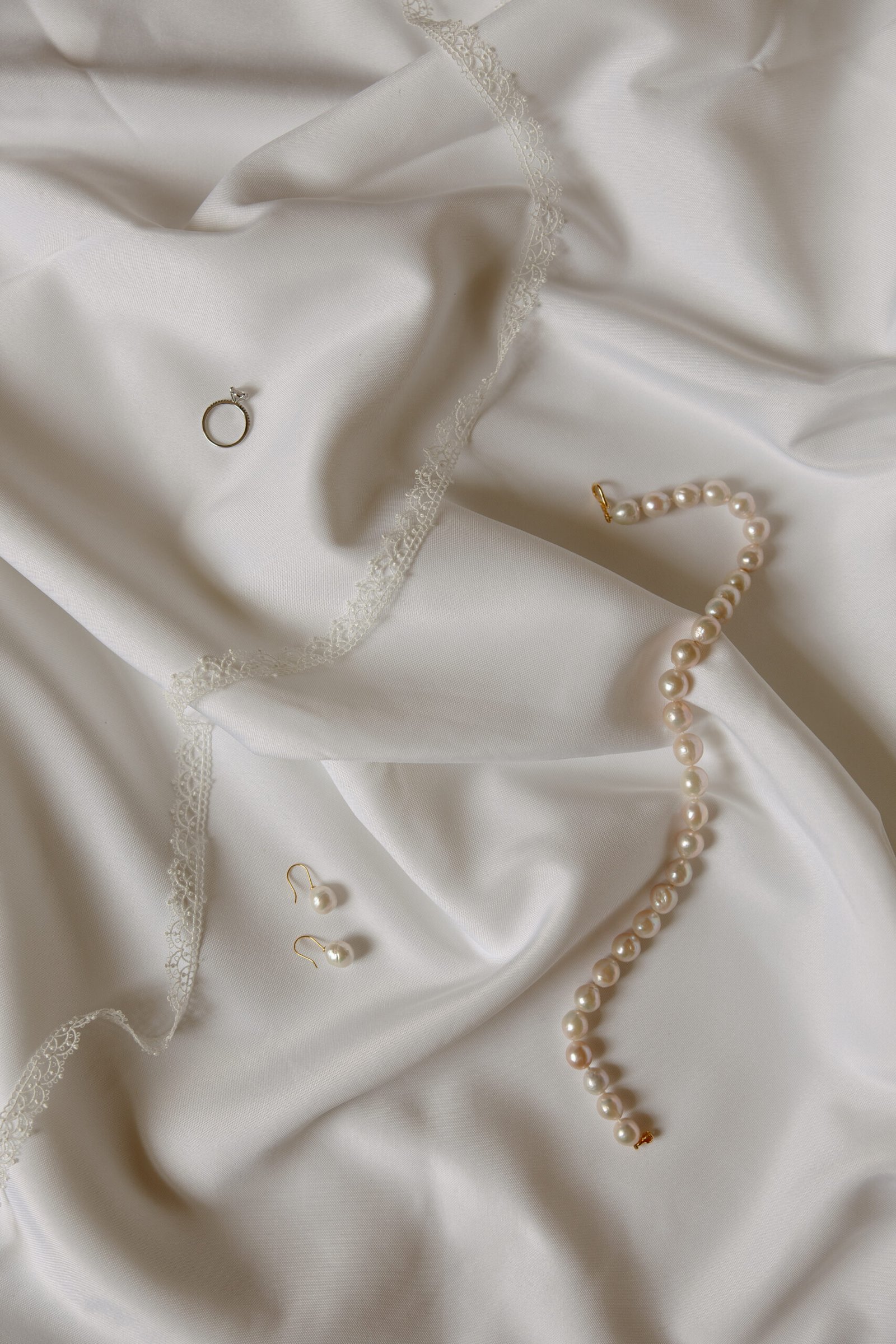 A diamond ring, a pearl bracelet, and a pair of pearl earrings are displayed on white satin fabric with lace trim.